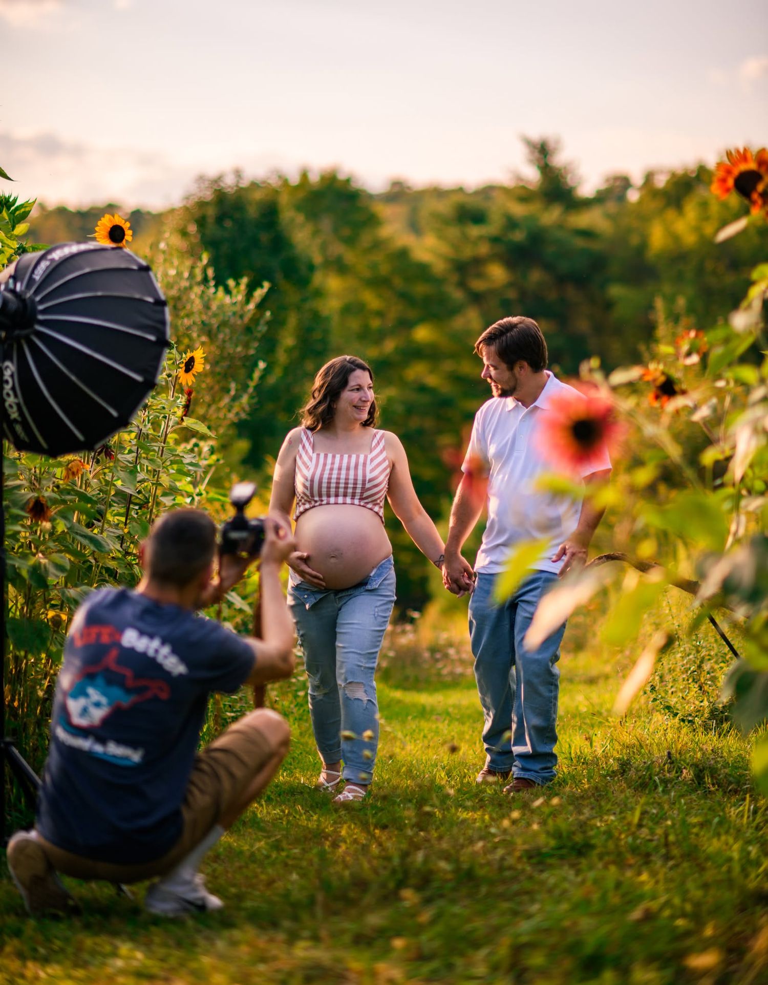 Photographer captures pregnant couple holding hands in sunflower field during golden hour maternity photo session outdoors.