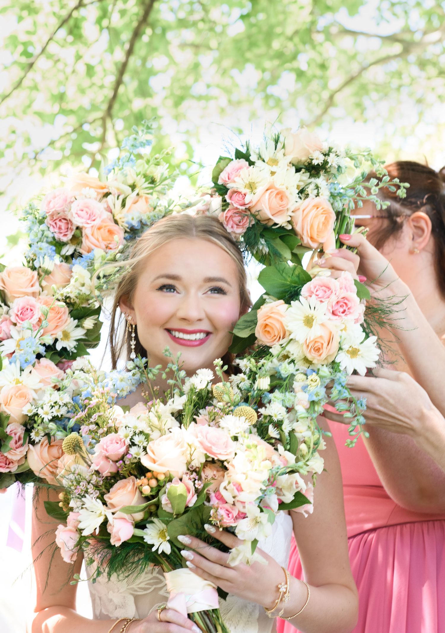 Smiling bride framed by pastel pink and peach floral bouquets at an outdoor wedding ceremony.
