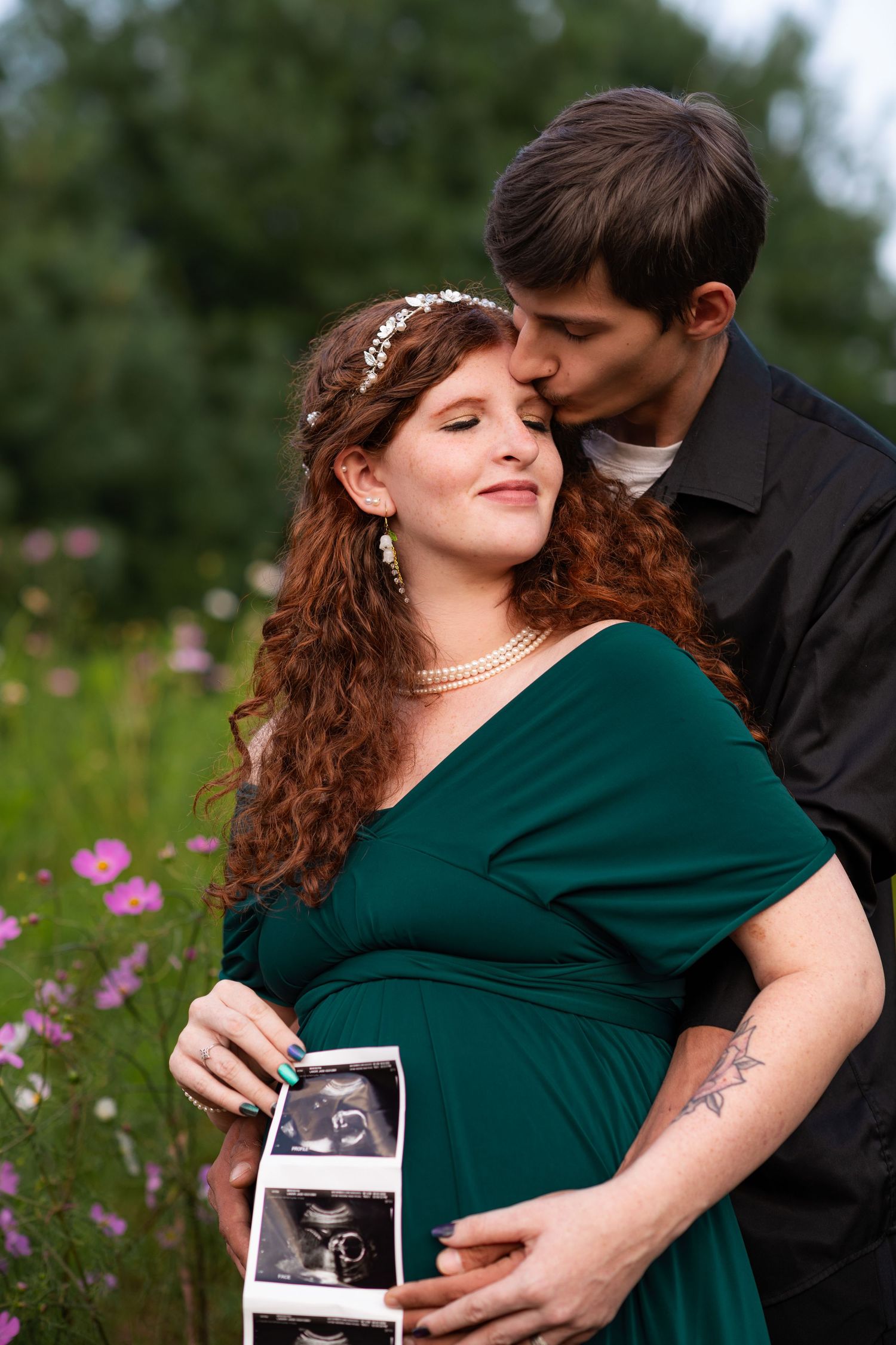 Pregnant woman in green dress holds ultrasound photos while partner kisses her cheek outdoors.