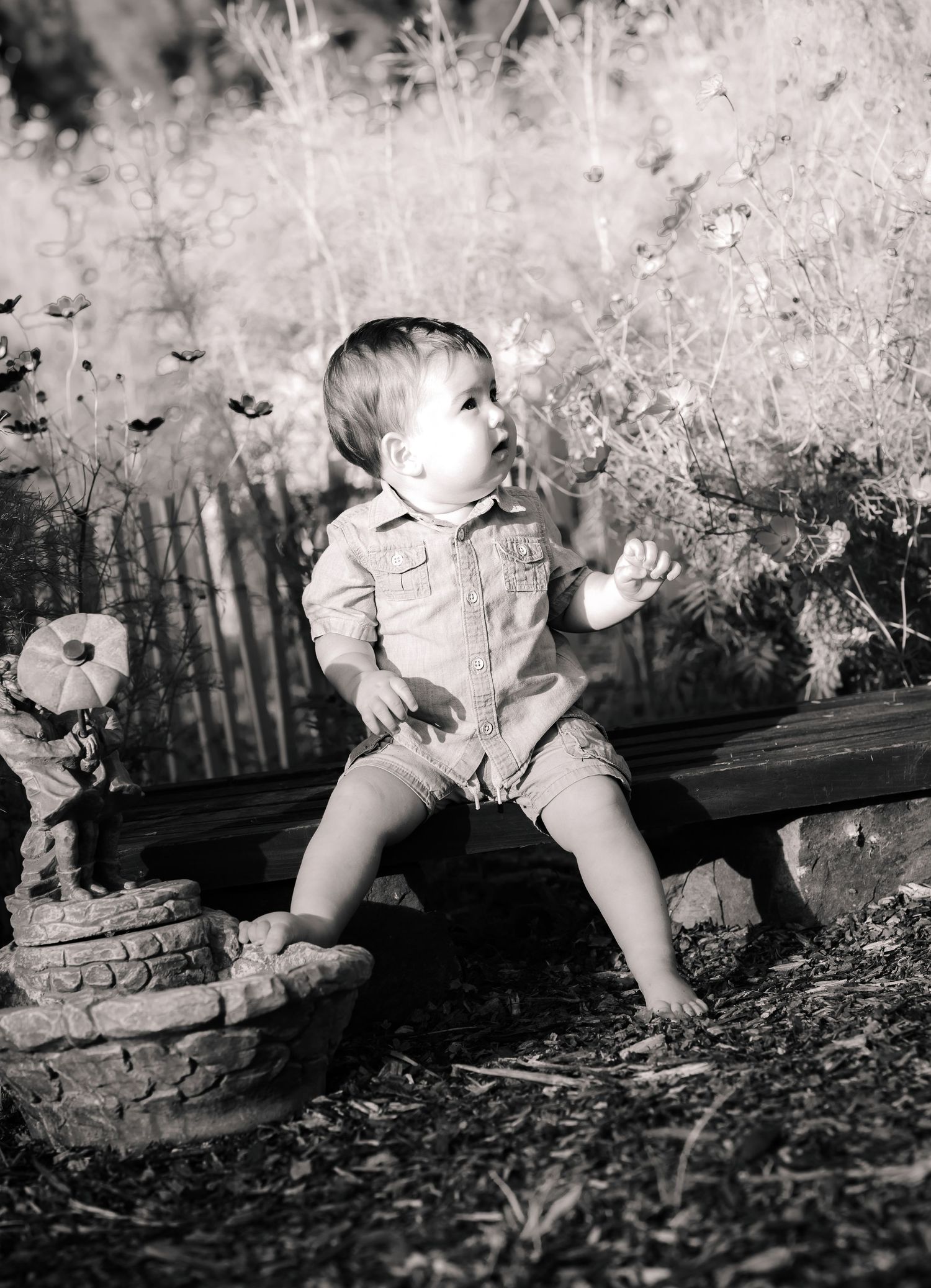 Black and white photo of a curious toddler sitting on a garden bench surrounded by flowers and plants.