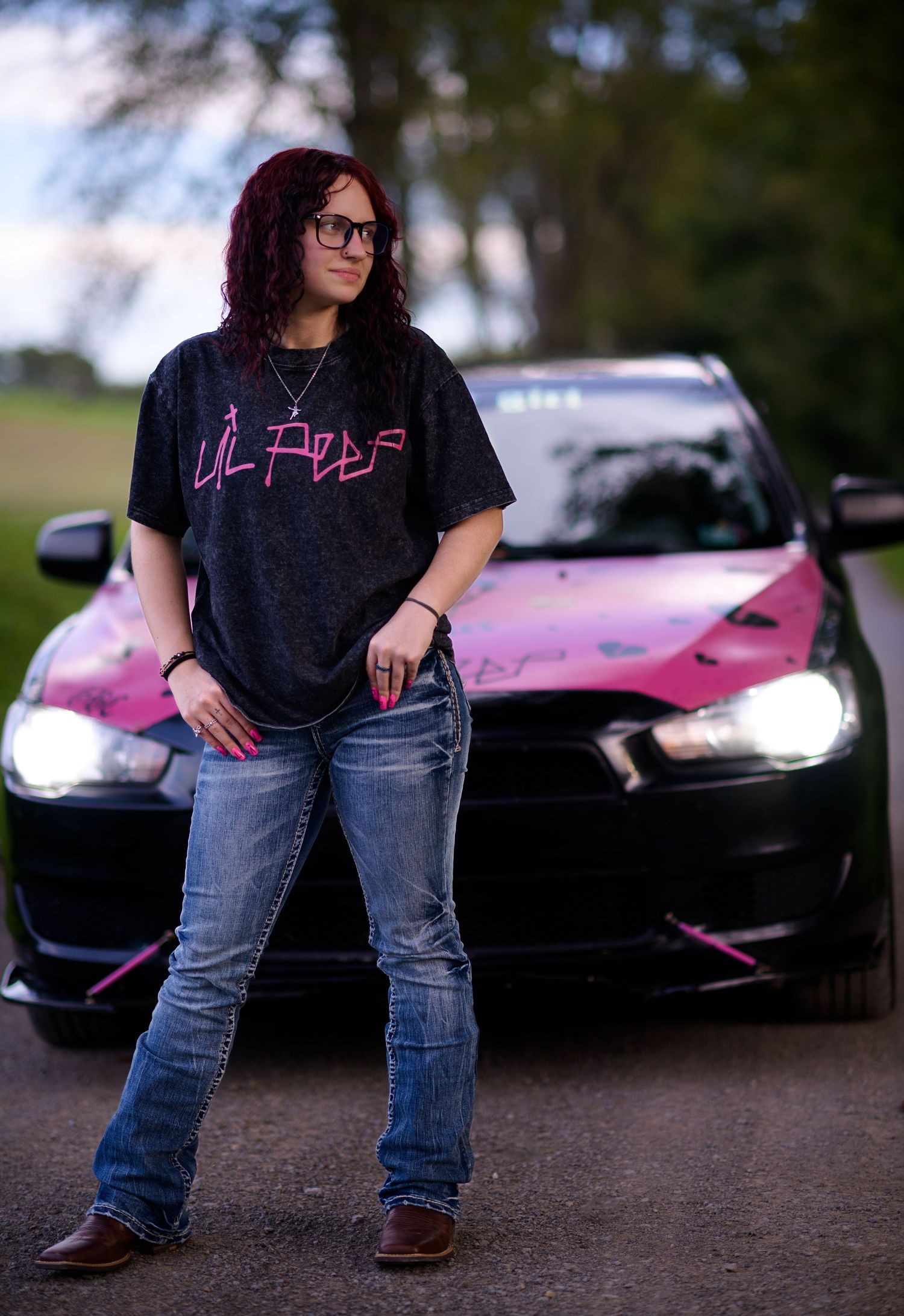 Young woman with red curly hair in black graphic tee and jeans poses beside pink and black wrapped car.