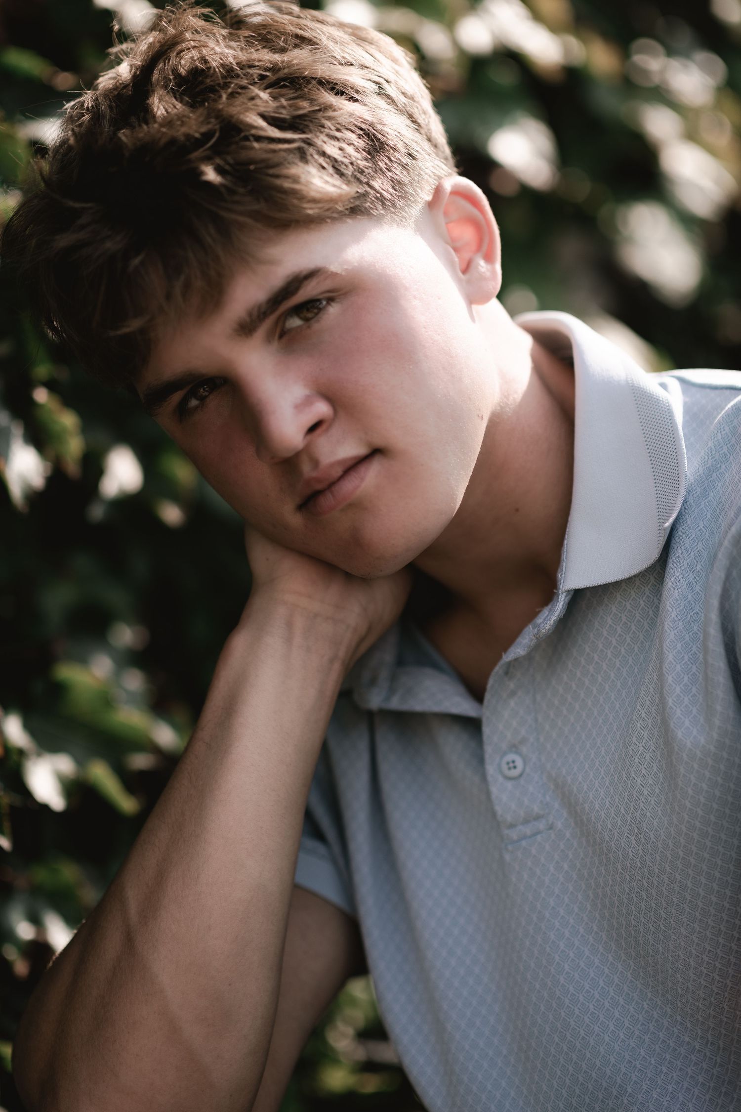 Young man in light blue polo shirt resting chin on hand, posed outdoors with green foliage background.