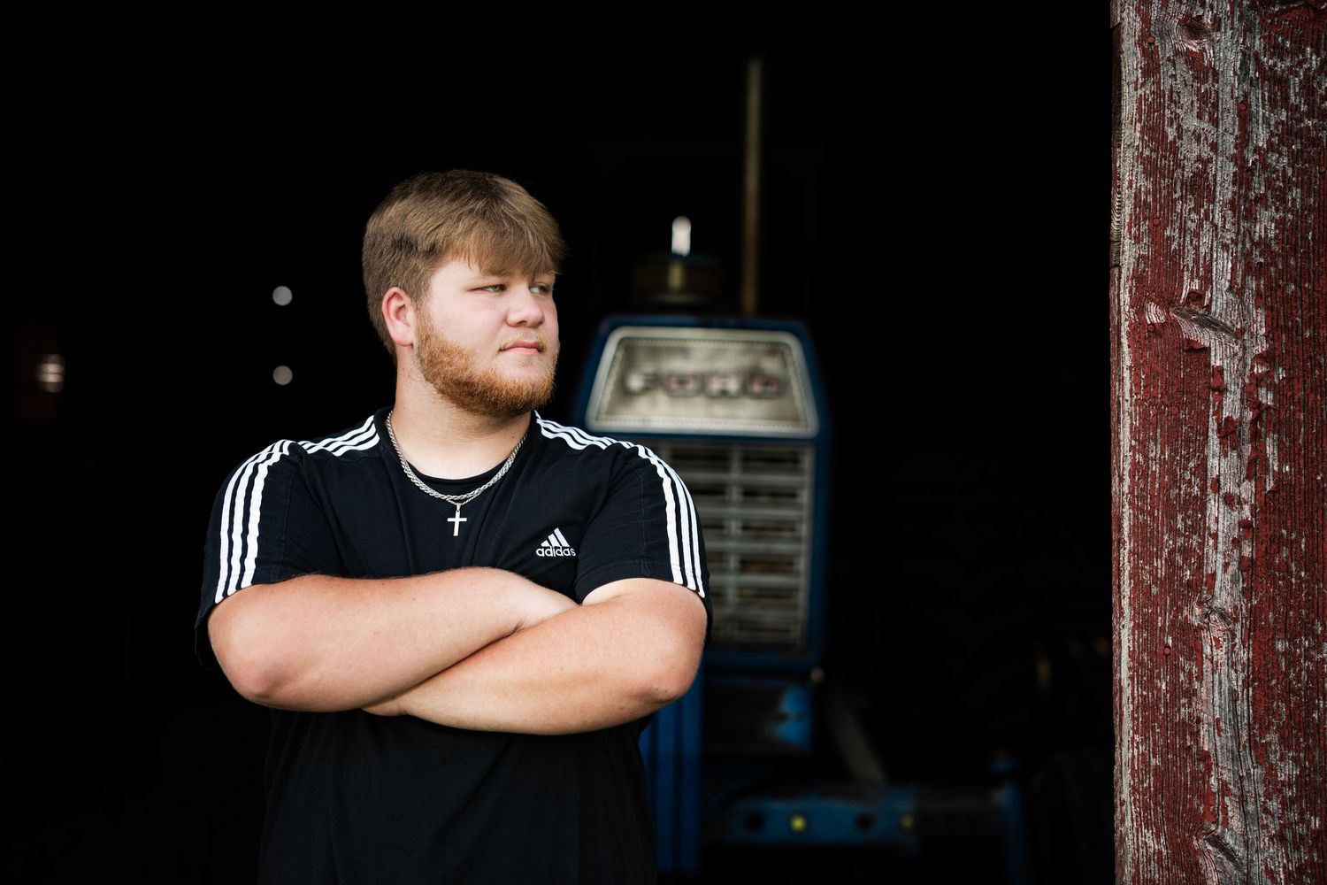 Young man in black Adidas shirt with cross necklace stands arms crossed outdoors near rustic red barn.