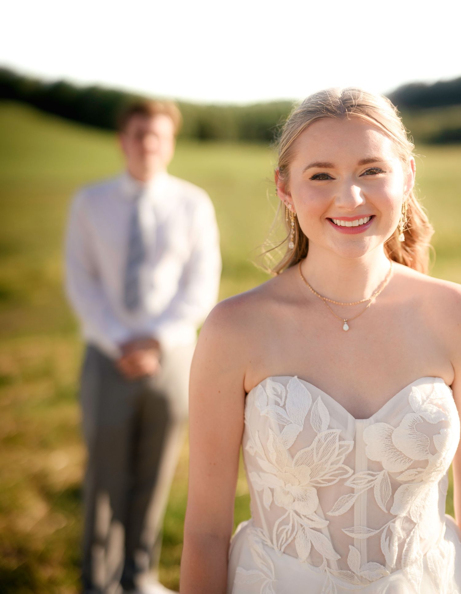 Smiling bride in strapless floral lace gown stands outdoors with groom in white shirt and tie blurred in background.