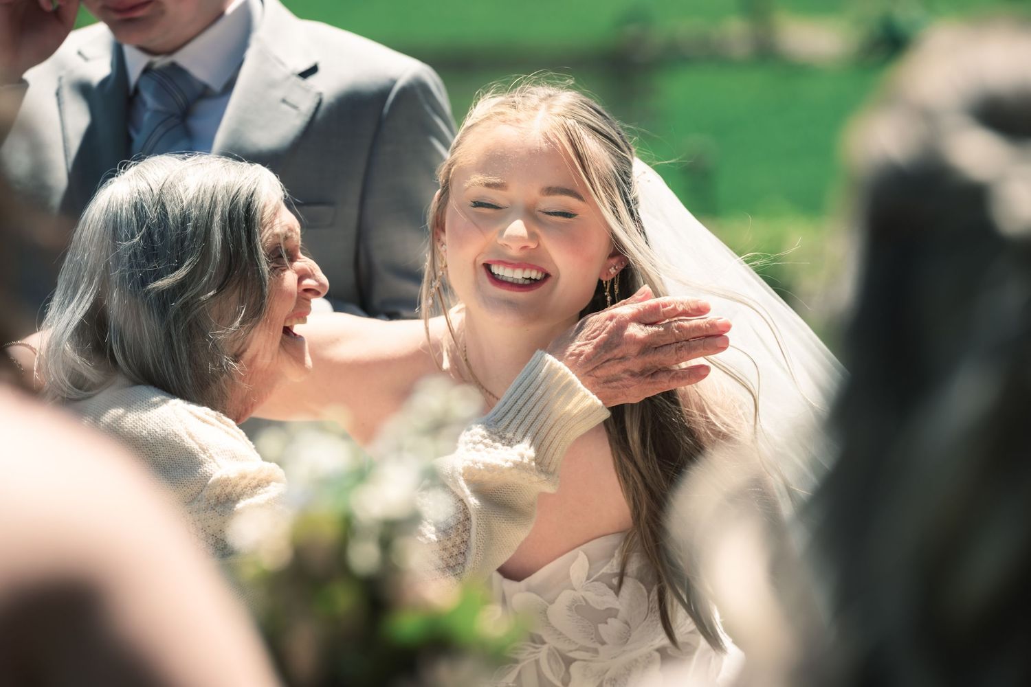 Bride in veil laughing joyfully with older woman at outdoor wedding ceremony.