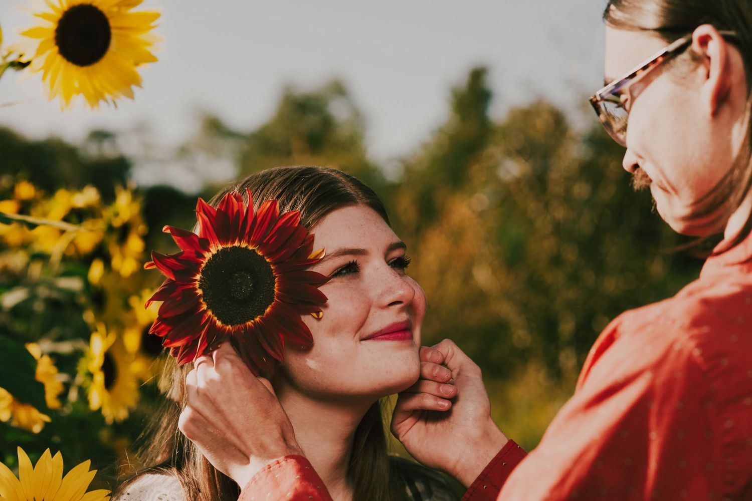 A woman smiles as another places a red sunflower behind her ear in a sunny garden.