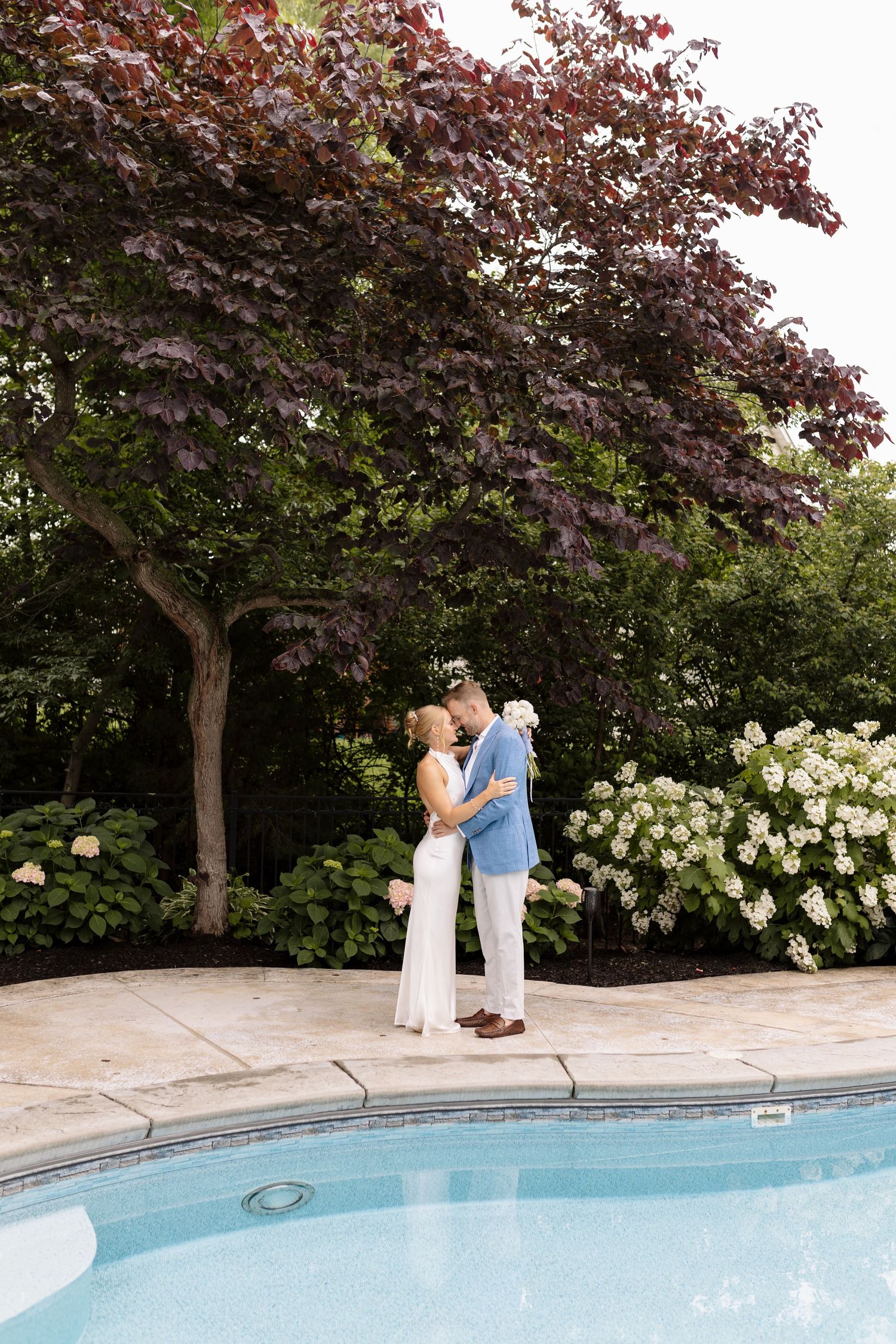 Couple embracing poolside with lush green trees and white hydrangeas in a romantic outdoor garden setting.