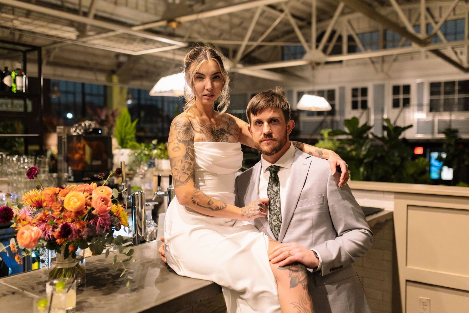 Tattooed bride in white dress and groom in gray suit pose at bar in industrial wedding venue.