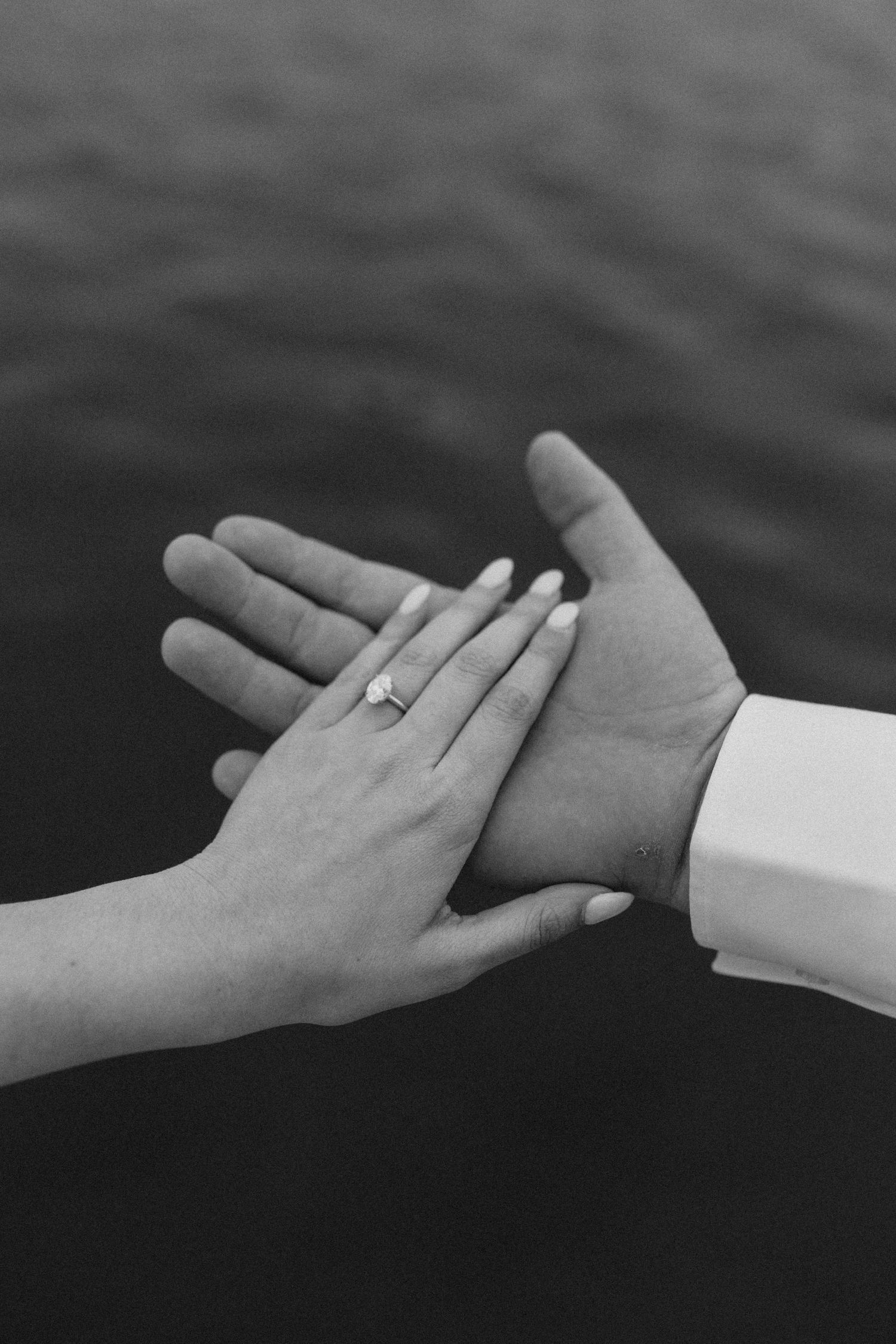 Black and white close-up of couple holding hands over water, woman wearing a diamond engagement ring.