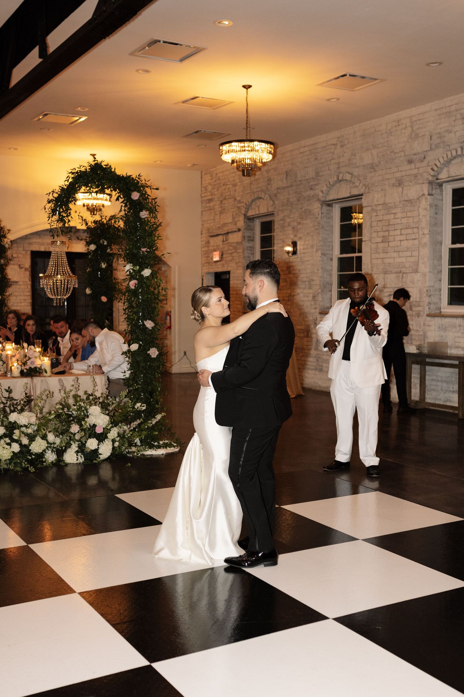 Bride and groom share first dance on checkered floor with violinist performing at elegant wedding reception.
