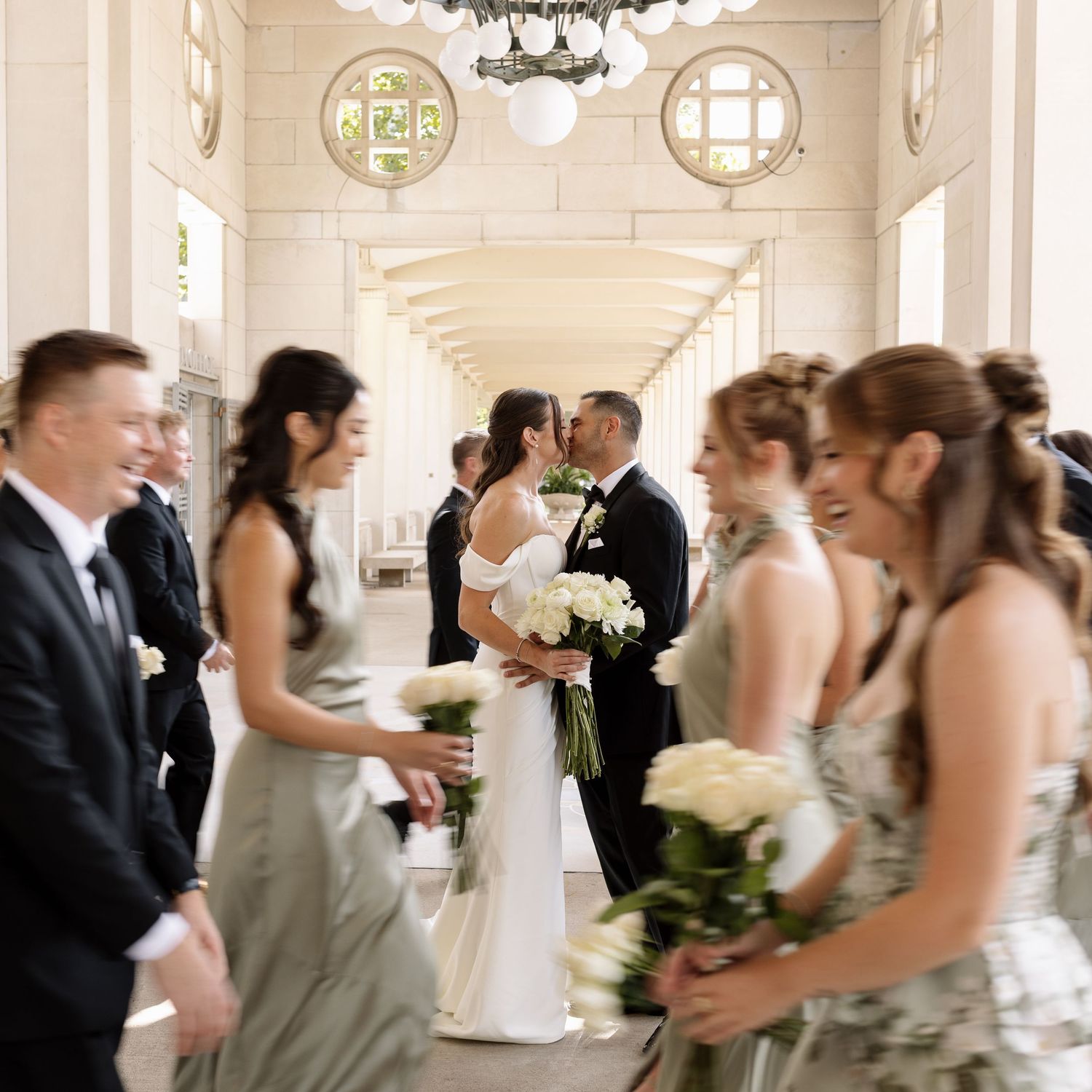 Bride and groom sharing a kiss while wedding party in sage gowns and black suits surrounds them indoors.