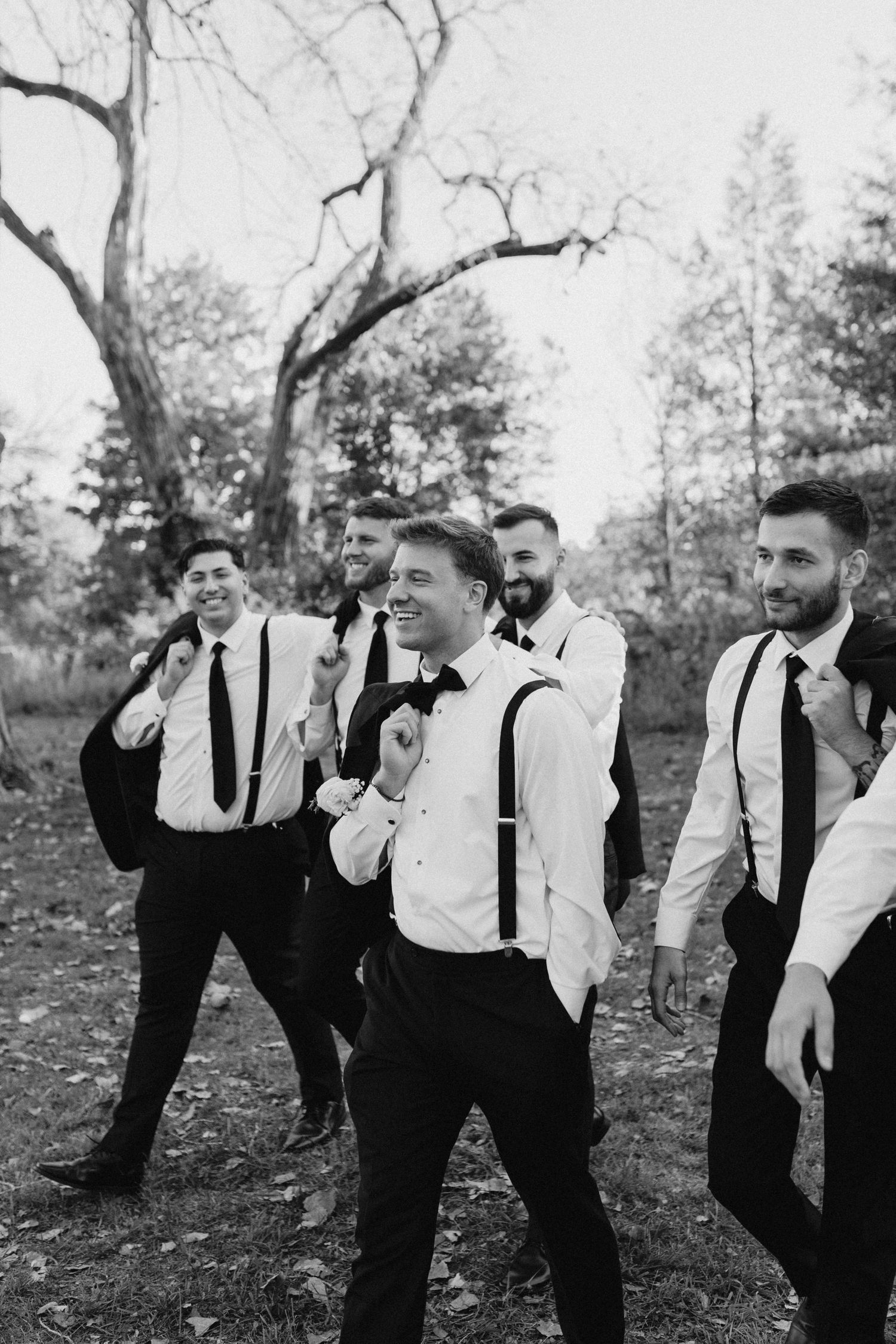 Black and white photo of groom and groomsmen in suspenders and ties walking outdoors at a wedding.