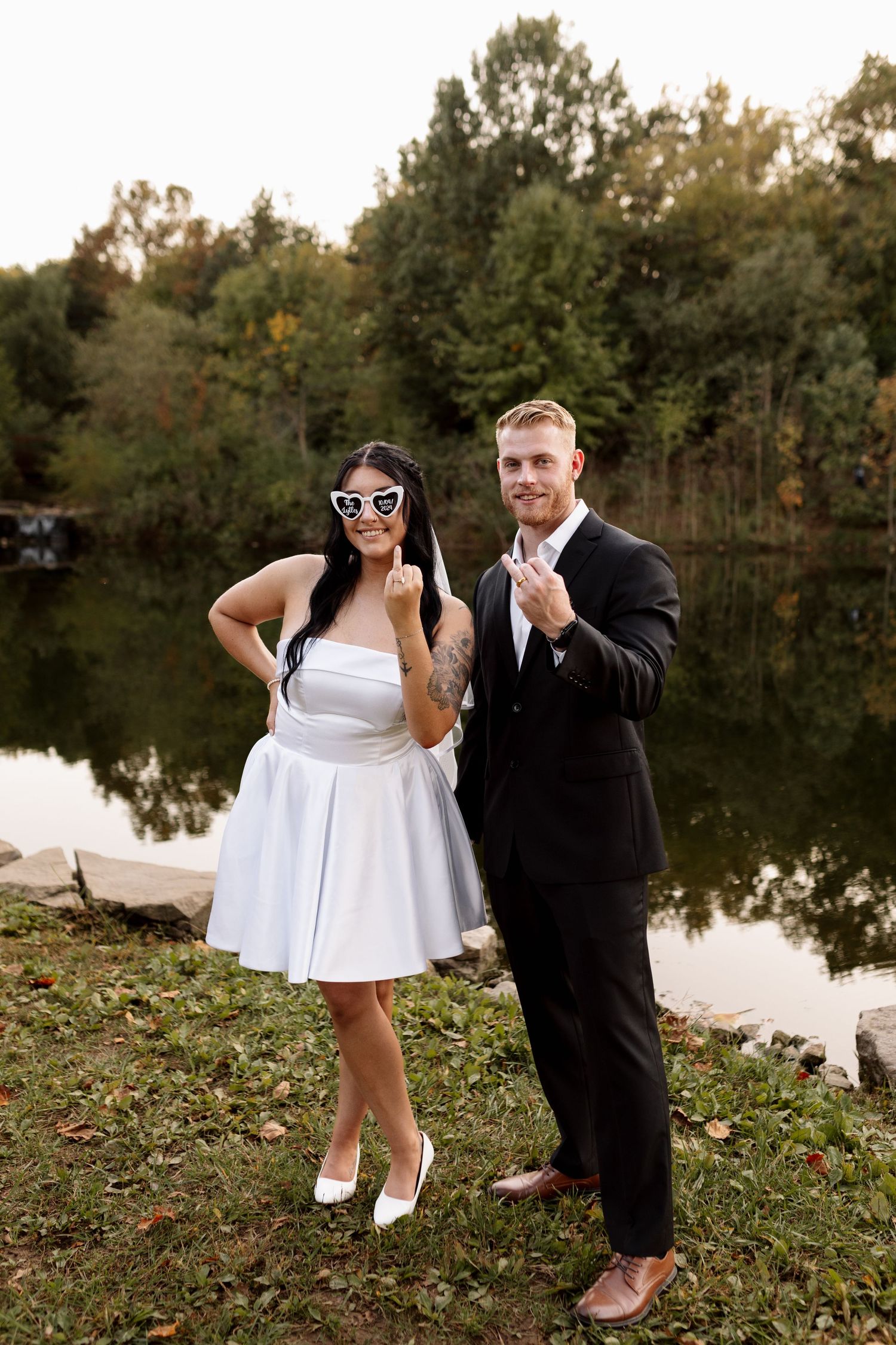Fun couple in wedding attire showing rings by a lake, bride wearing heart sunglasses.