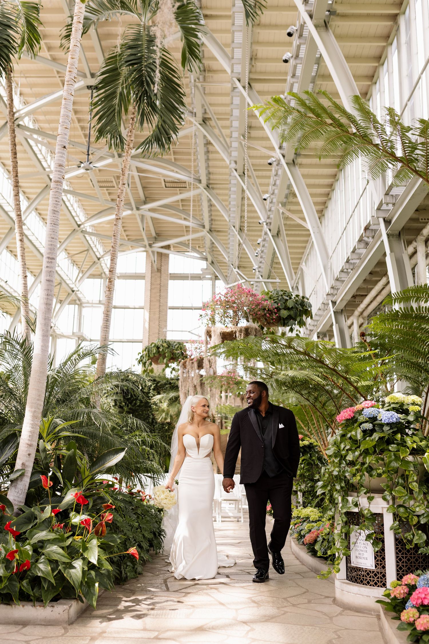 Bride and groom walking hand in hand through a lush indoor botanical garden atrium on their wedding day.