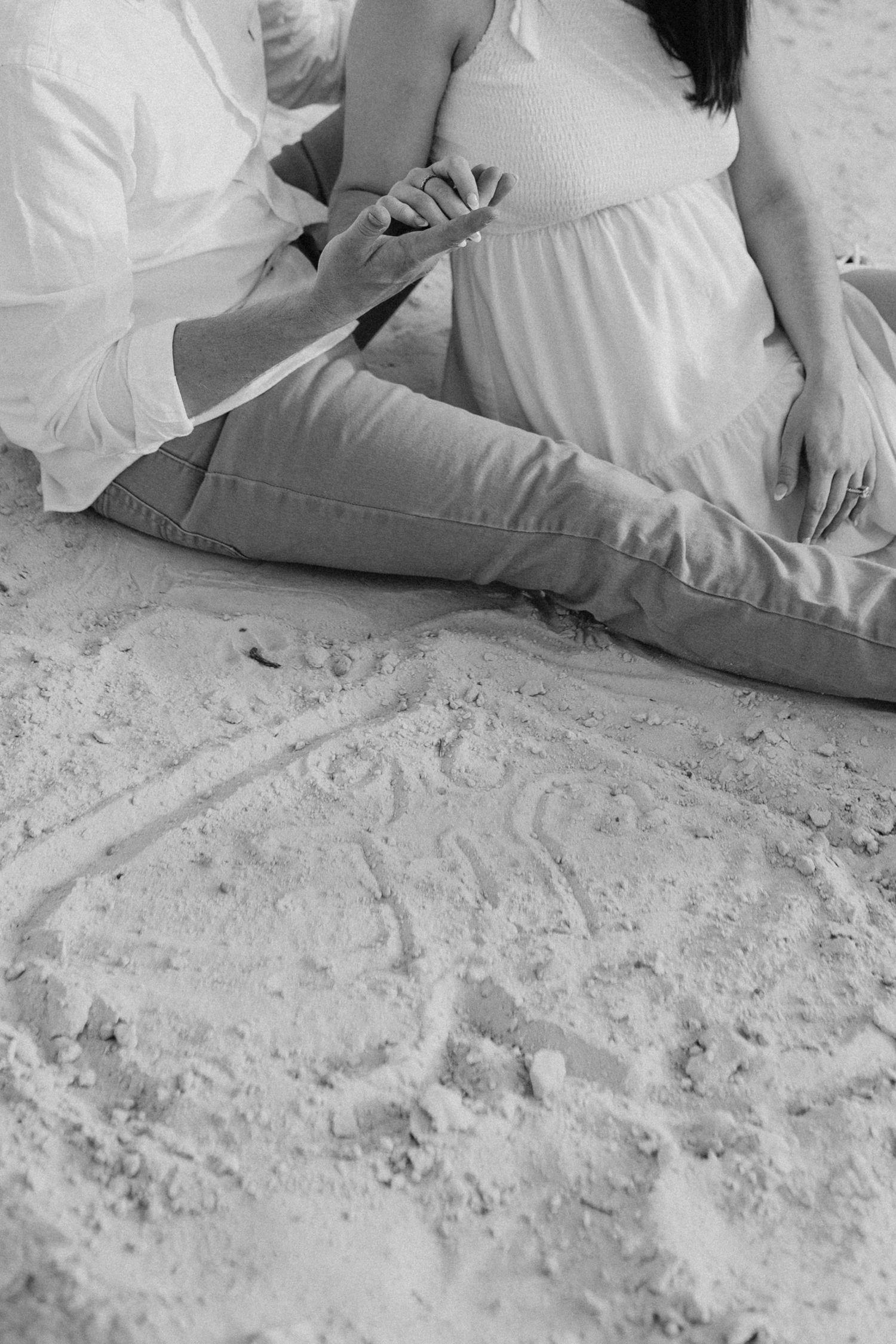 Black and white photo of couple sitting on sandy beach holding hands with heart drawn in sand foreground.