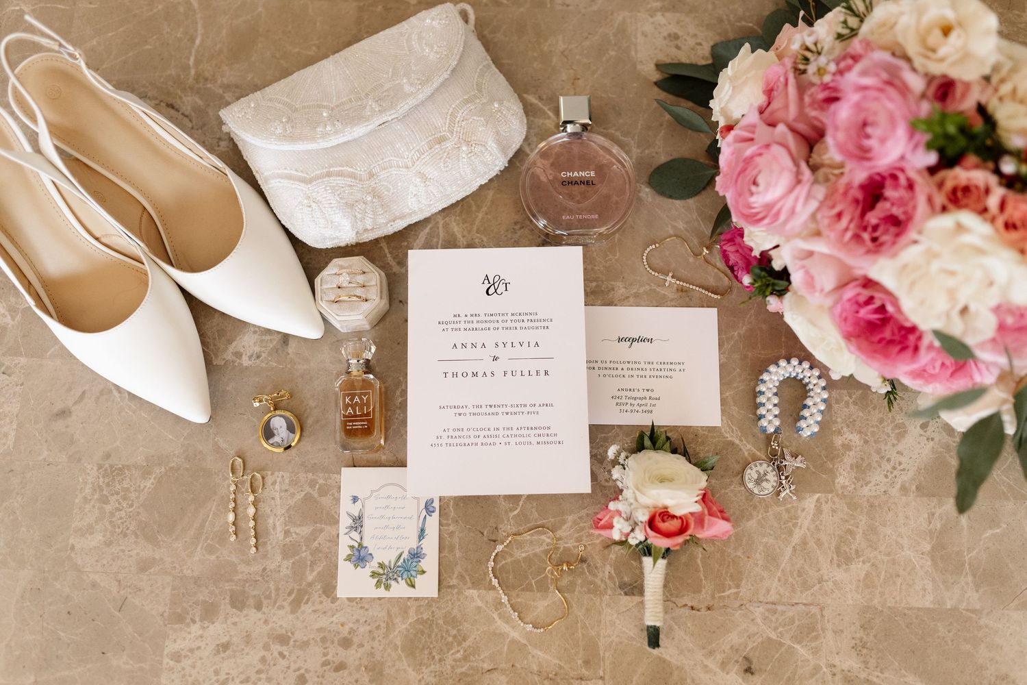 Elegant wedding flat lay with invitation suite, white heels, pink bouquet, perfume bottles, and delicate jewelry on marble.