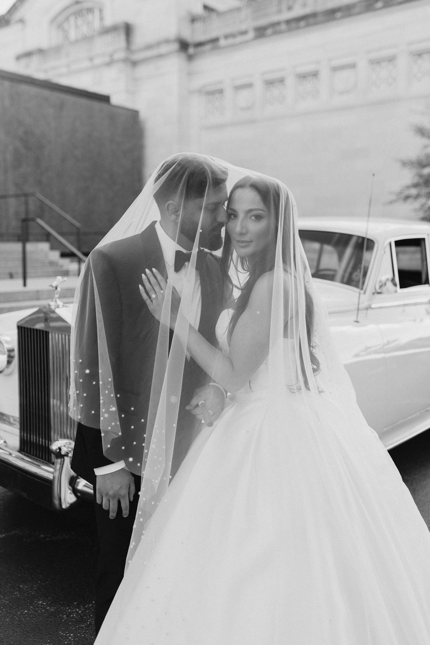 Bride and groom sharing intimate moment under pearl-adorned veil beside classic white Rolls-Royce.