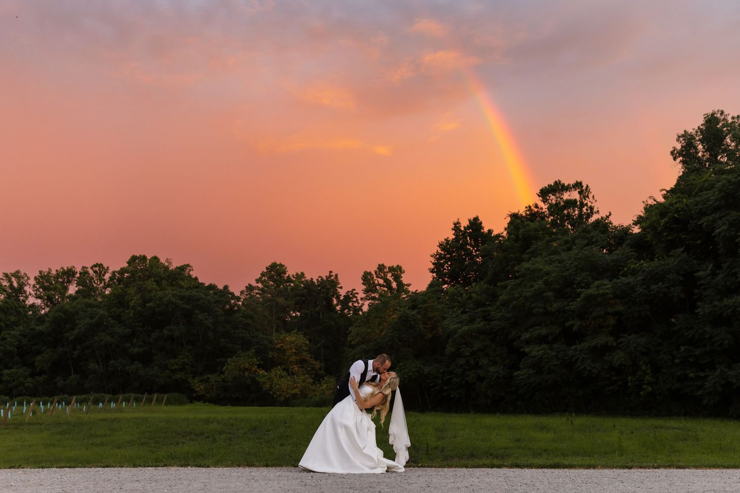 Bride and groom kissing at sunset with a stunning rainbow arching over lush trees.