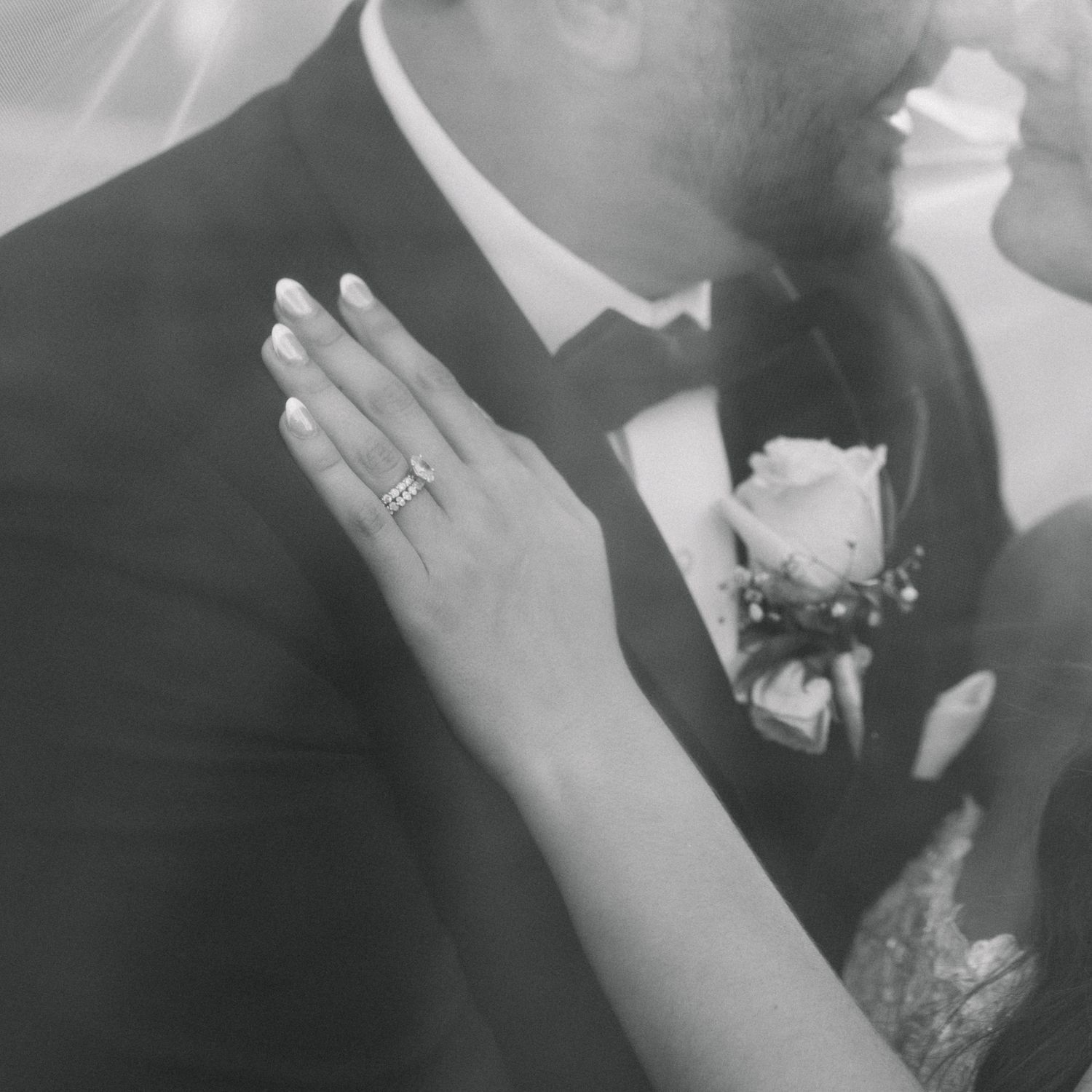 Black and white close-up of bride's hand with diamond ring resting on groom's tuxedo lapel.