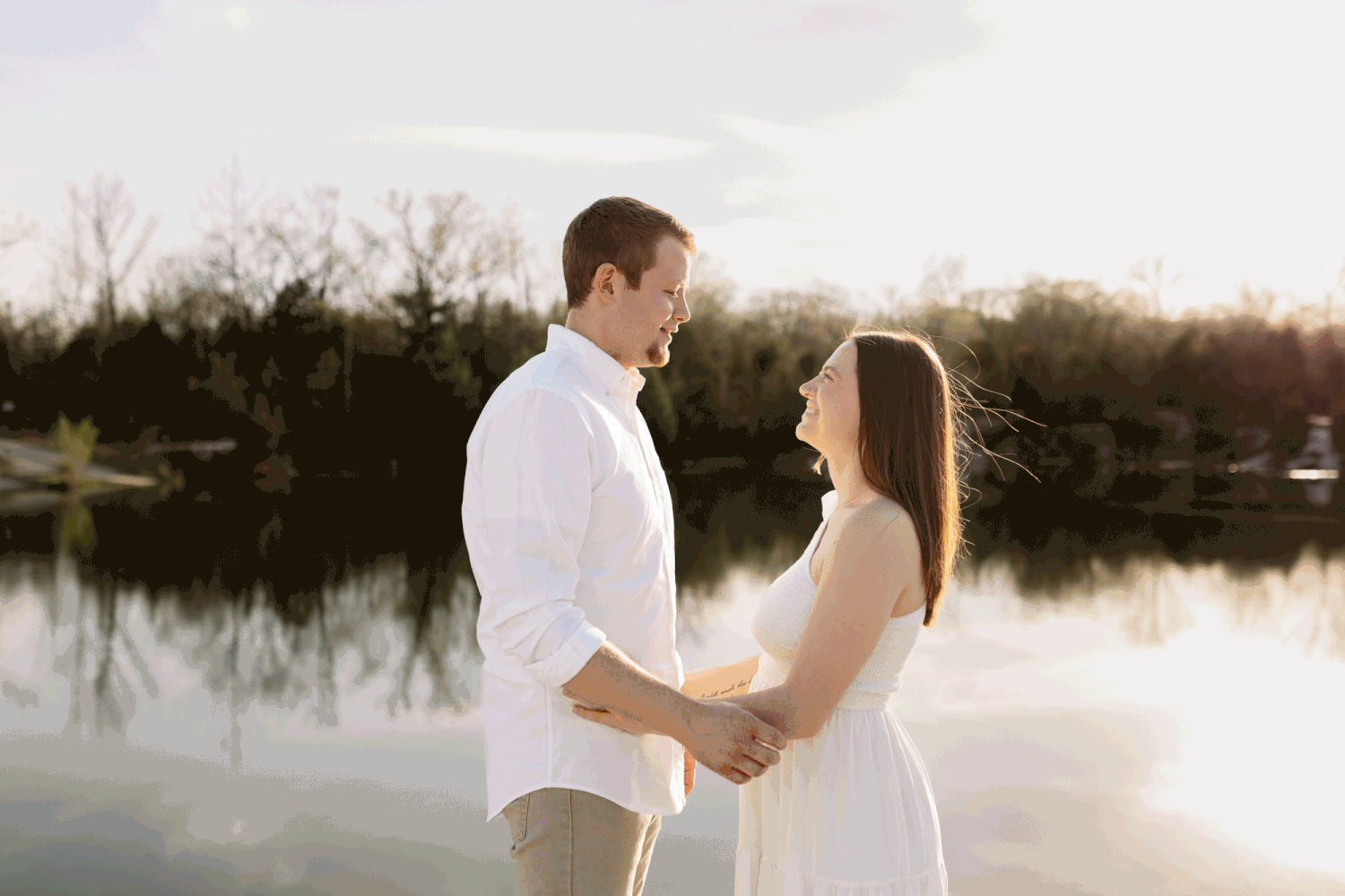 A gif of a couple sharing a kiss in front of a lake surrounded by trees and sand on a windy day.