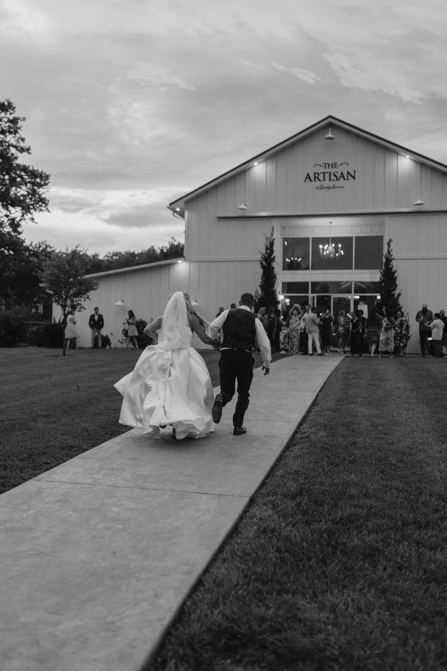 Black and white photo of bride and groom walking hand in hand toward The Artisan wedding venue at dusk.