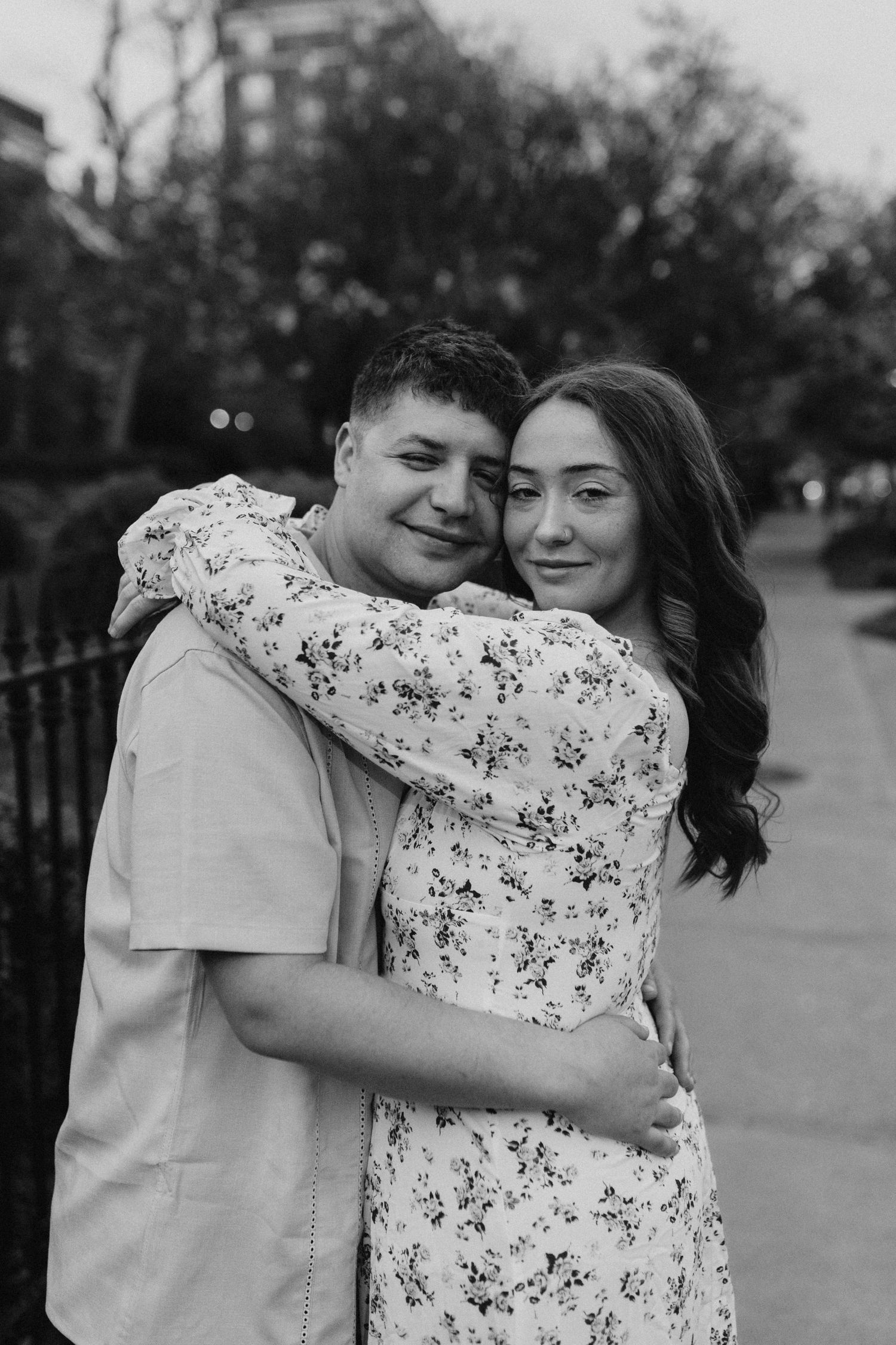 Black and white portrait of a couple hugging on a city sidewalk, woman in floral dress embracing man warmly.