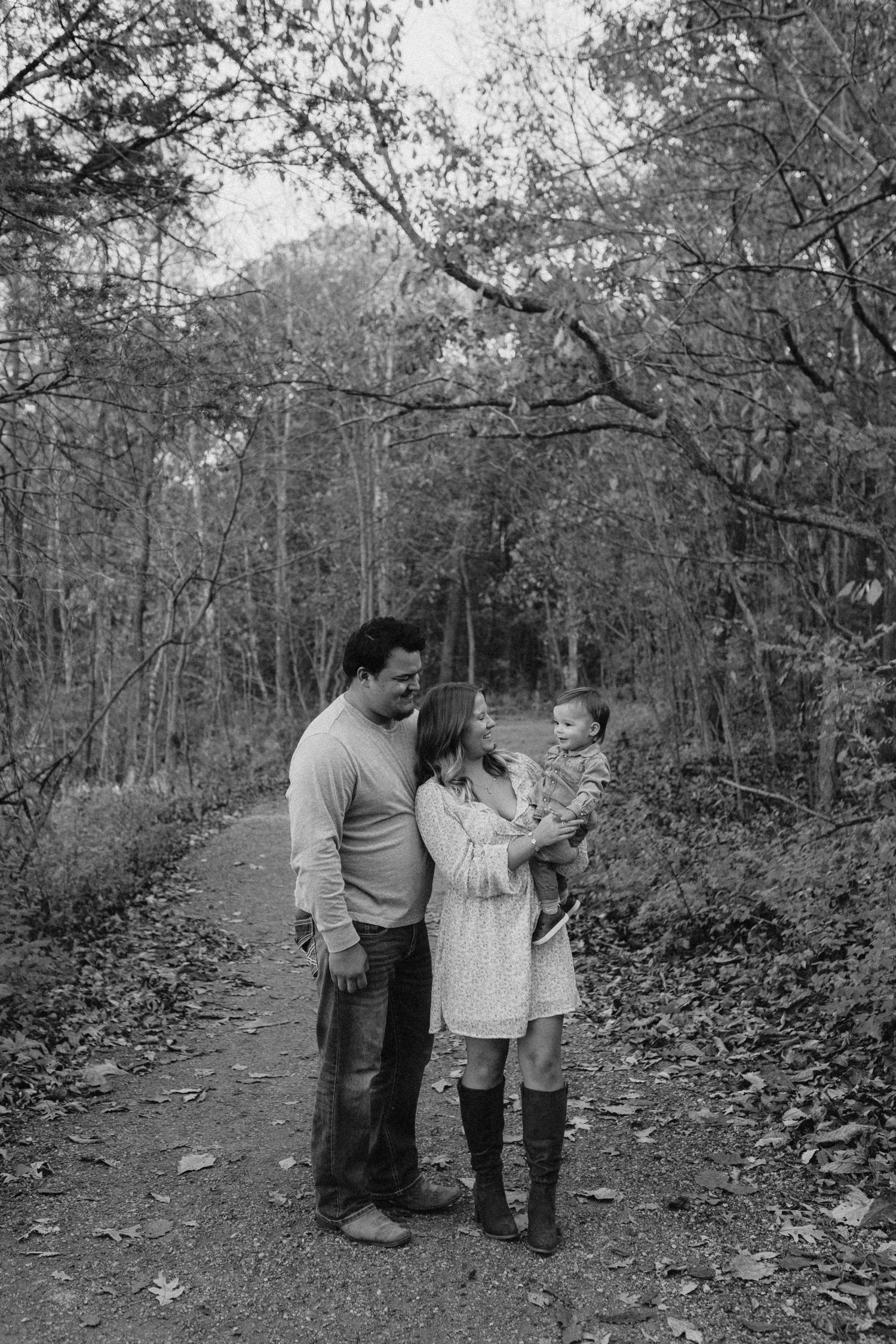 Black and white family portrait of parents with baby on a wooded forest trail in autumn.