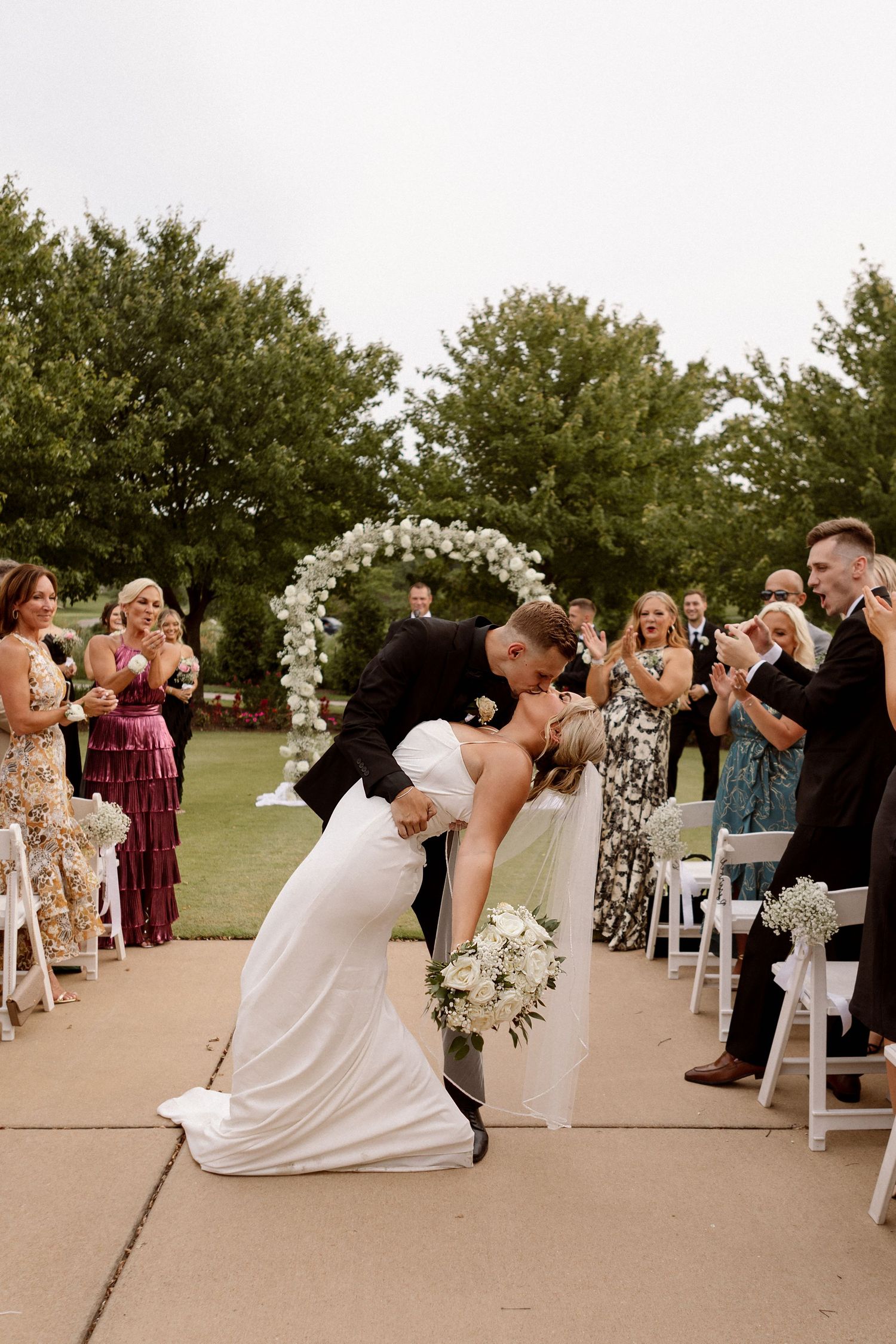 Groom dips bride for a kiss at outdoor wedding ceremony while guests applaud, with floral arch and green trees in background.