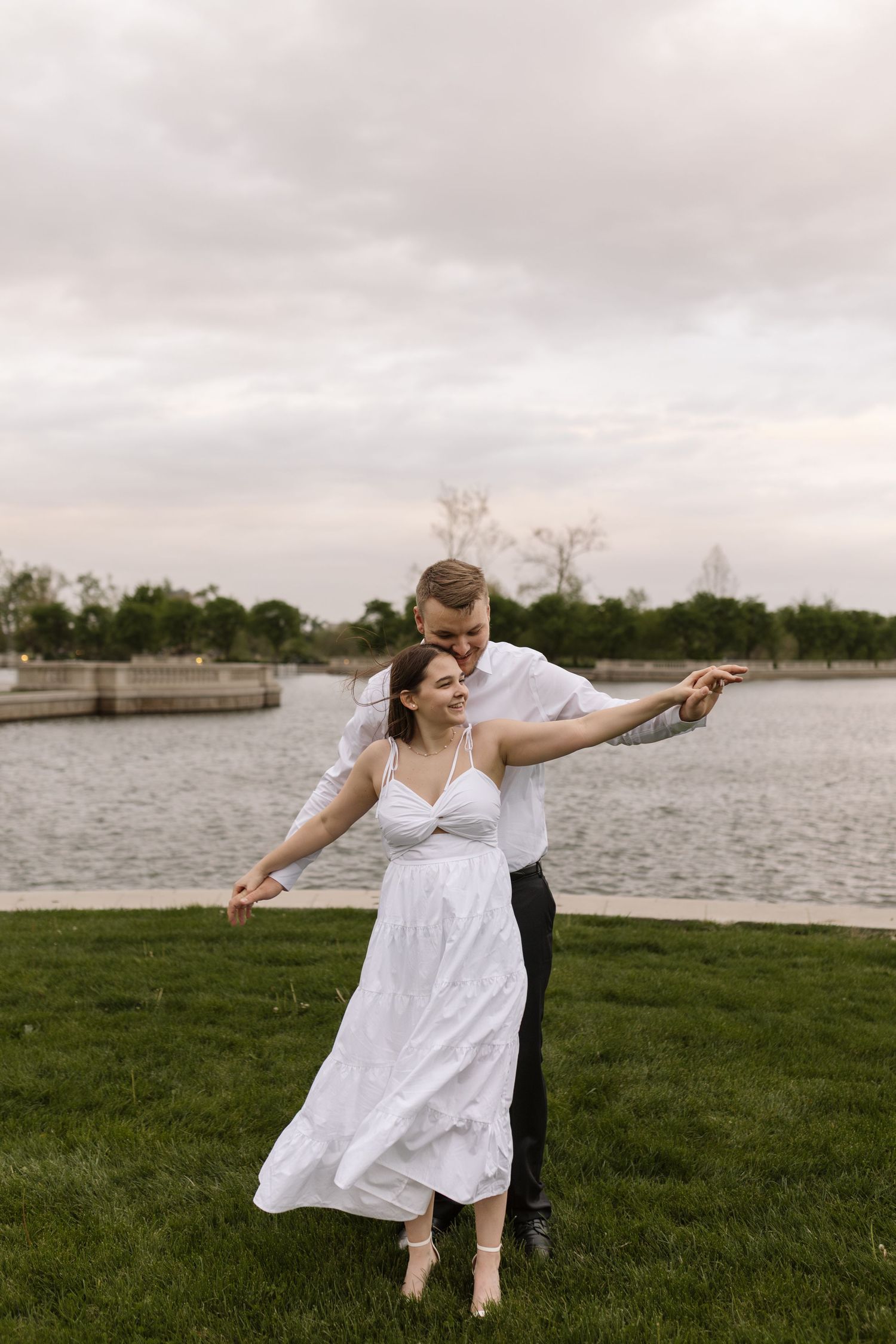 Couple dancing joyfully on green grass by a waterfront, groom in white shirt embracing bride in flowing dress.