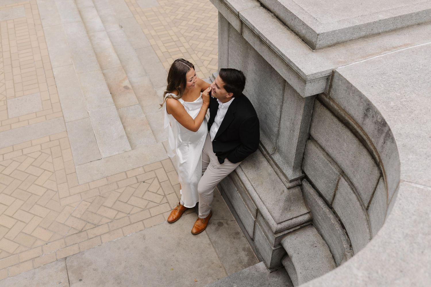 Couple sharing a romantic moment on grand stone steps, woman in white dress and man in black suit, aerial view.