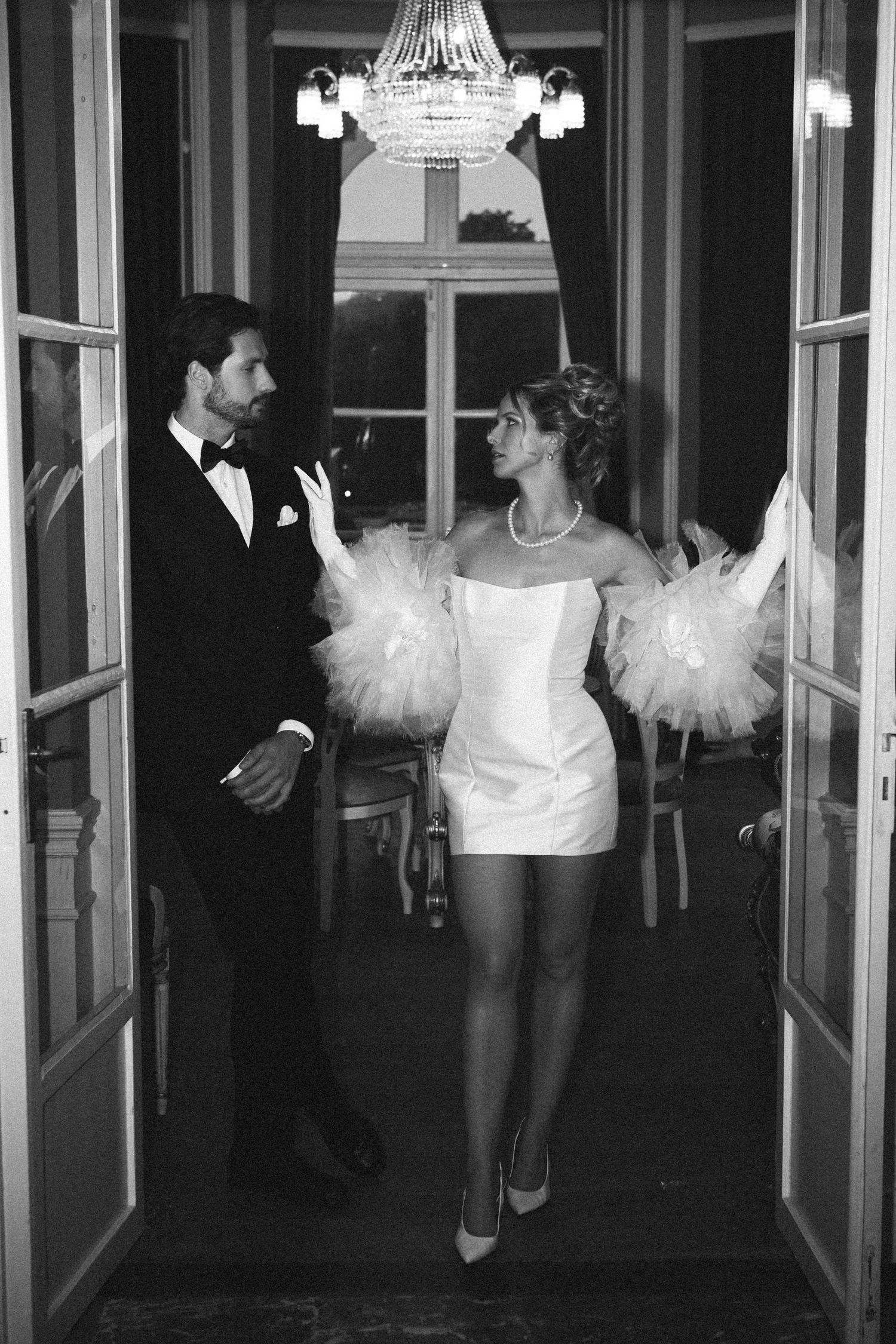 Black and white photo of a couple in wedding attire standing in doorway under chandelier, bride in short white dress.