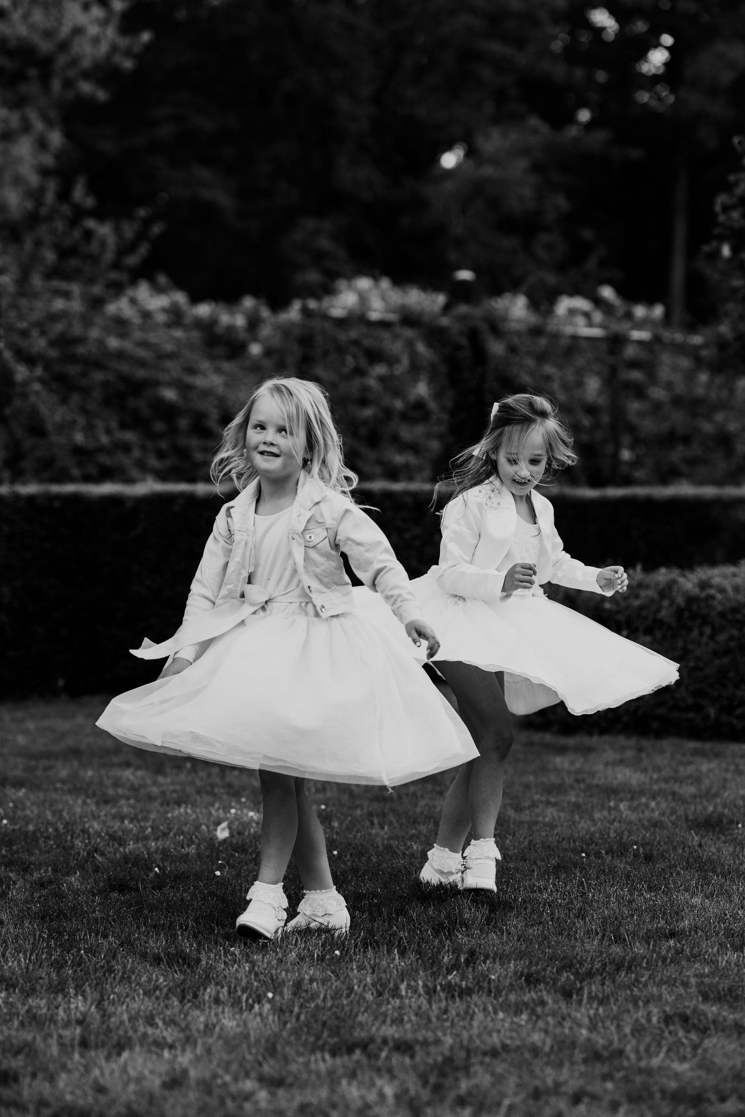 Two little flower girls in white dresses twirl and spin on a green lawn.