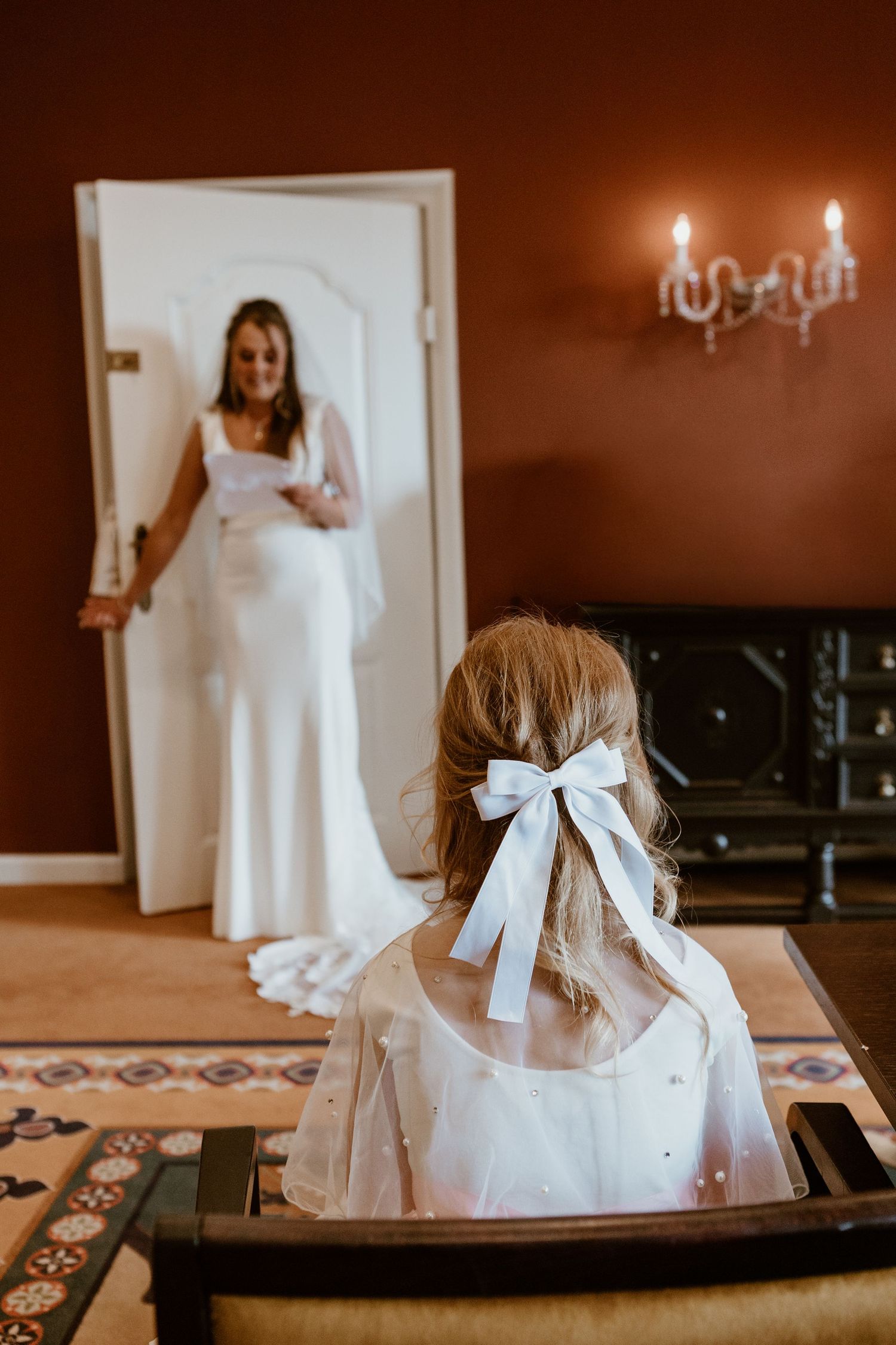 Flower girl with white bow in hair seated in chair as smiling bride in white gown appears in doorway.