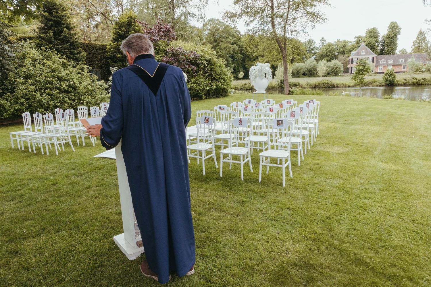 Officiant in blue robe stands at outdoor wedding altar with white chairs arranged on green lawn.