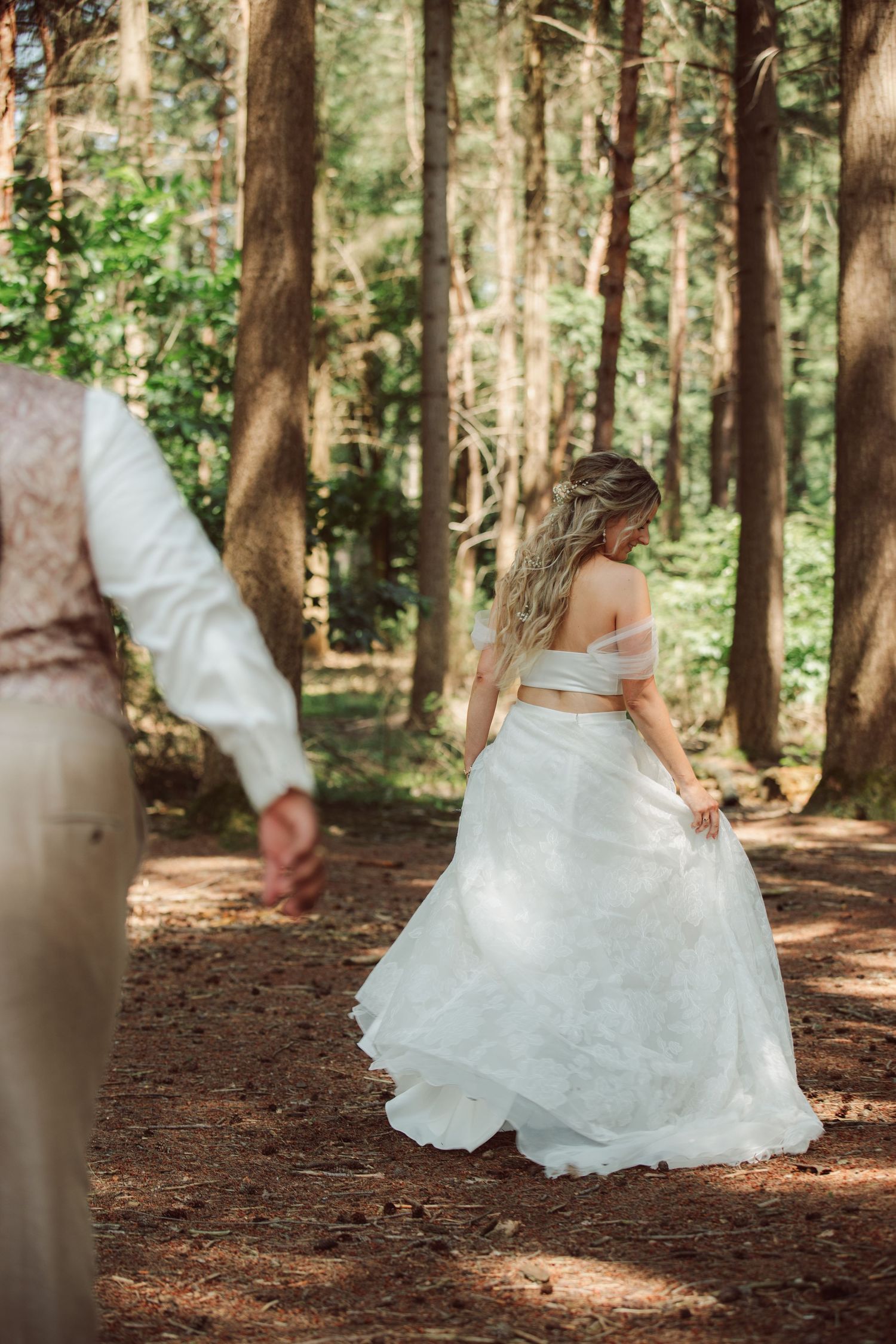 Bride in white gown walking through a sunlit forest with groom.