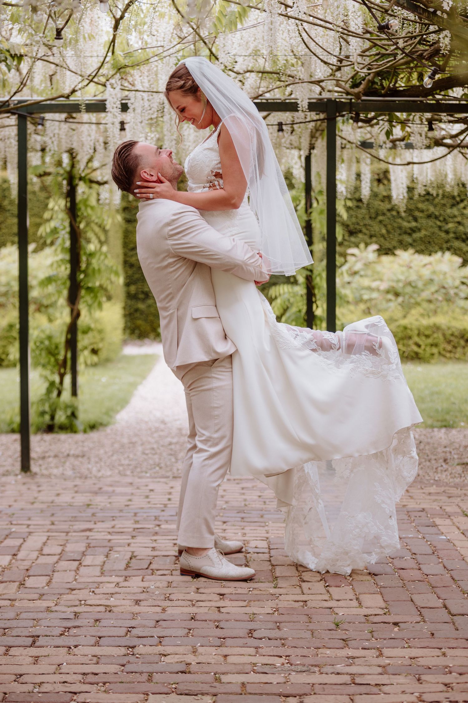 Groom lifts bride in white lace gown under blooming wisteria pergola in garden setting.