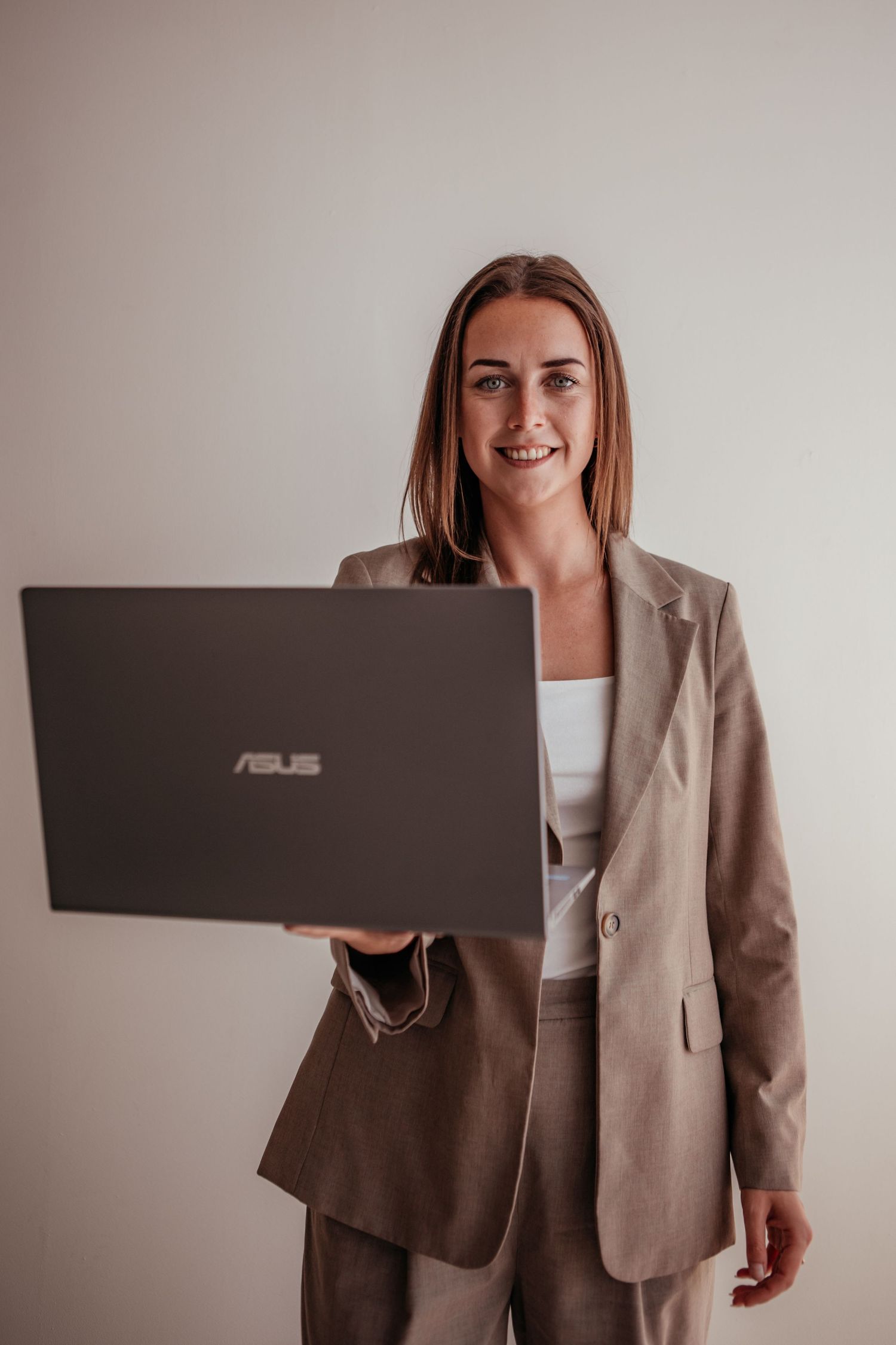 Smiling businesswoman in a beige suit holding an ASUS laptop against a light background.