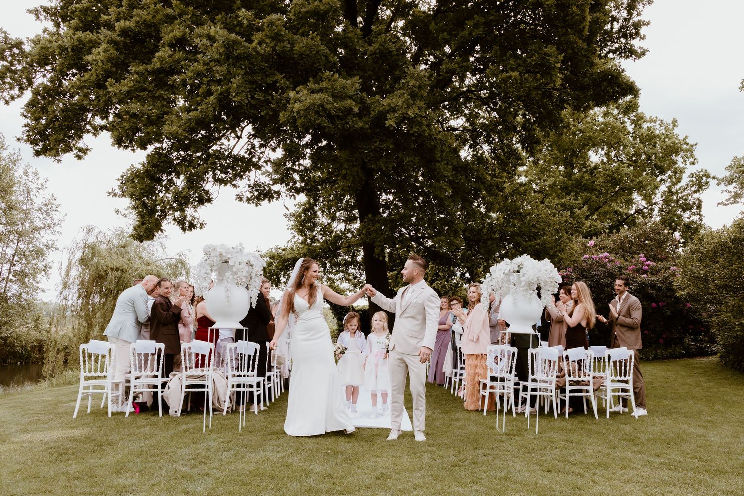 Wedding party celebrates outdoors on green lawn with white chairs and large tree backdrop.