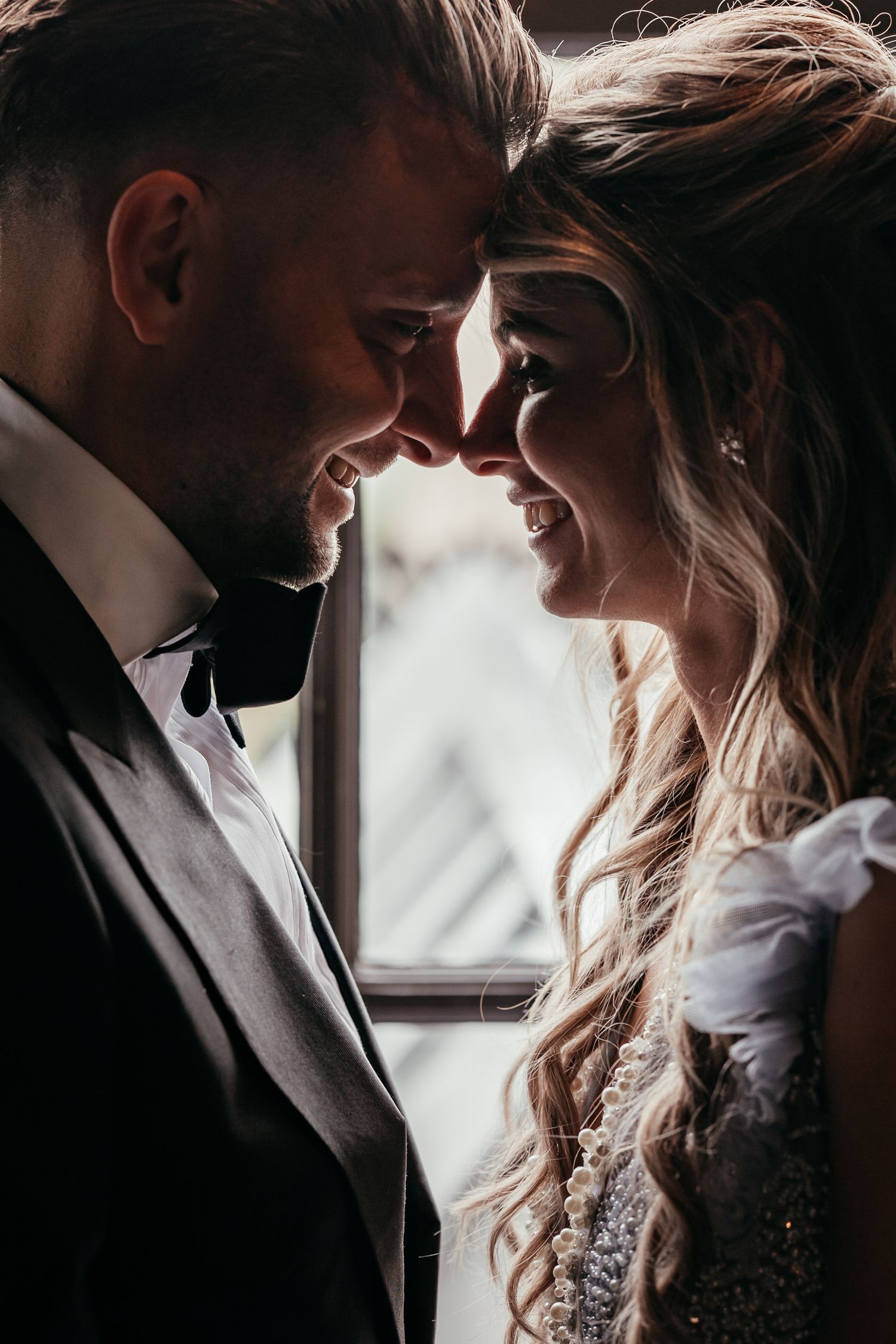 Romantic wedding couple touching foreheads and smiling in soft backlit window light.