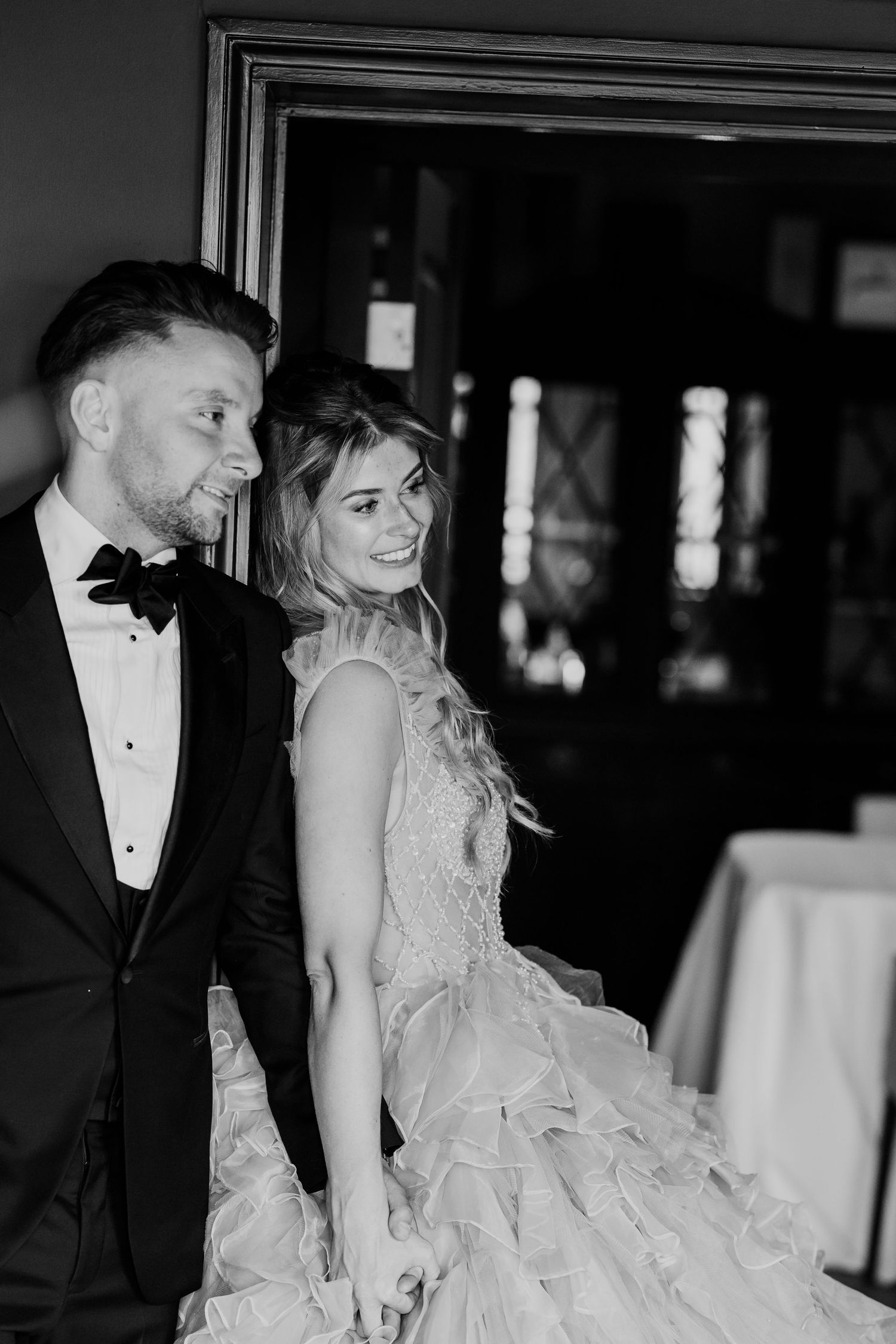 Black and white photo of smiling bride in ruffled gown holding hands with groom in tuxedo.