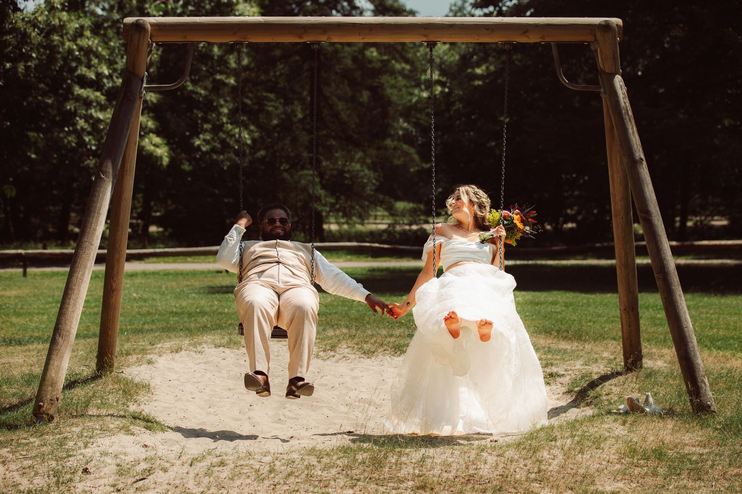 Bride and groom hold hands while sitting on swings at sunny outdoor wedding.