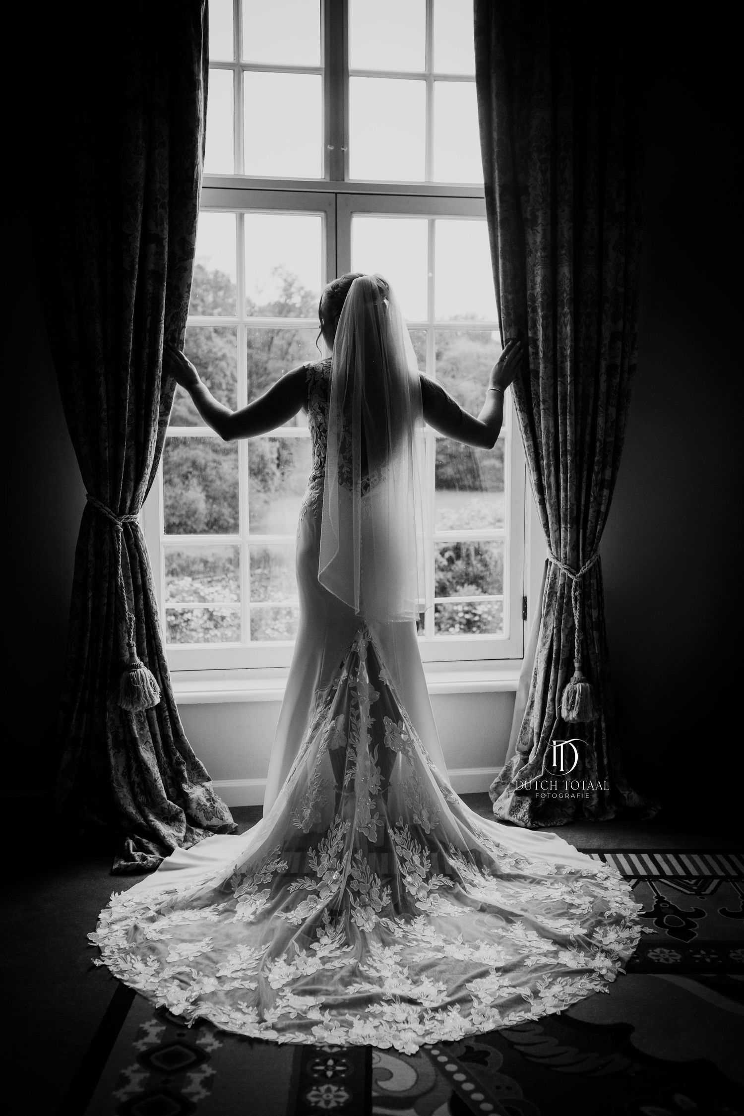 Dramatic black and white silhouette of bride in lace mermaid gown with long veil standing at large window.