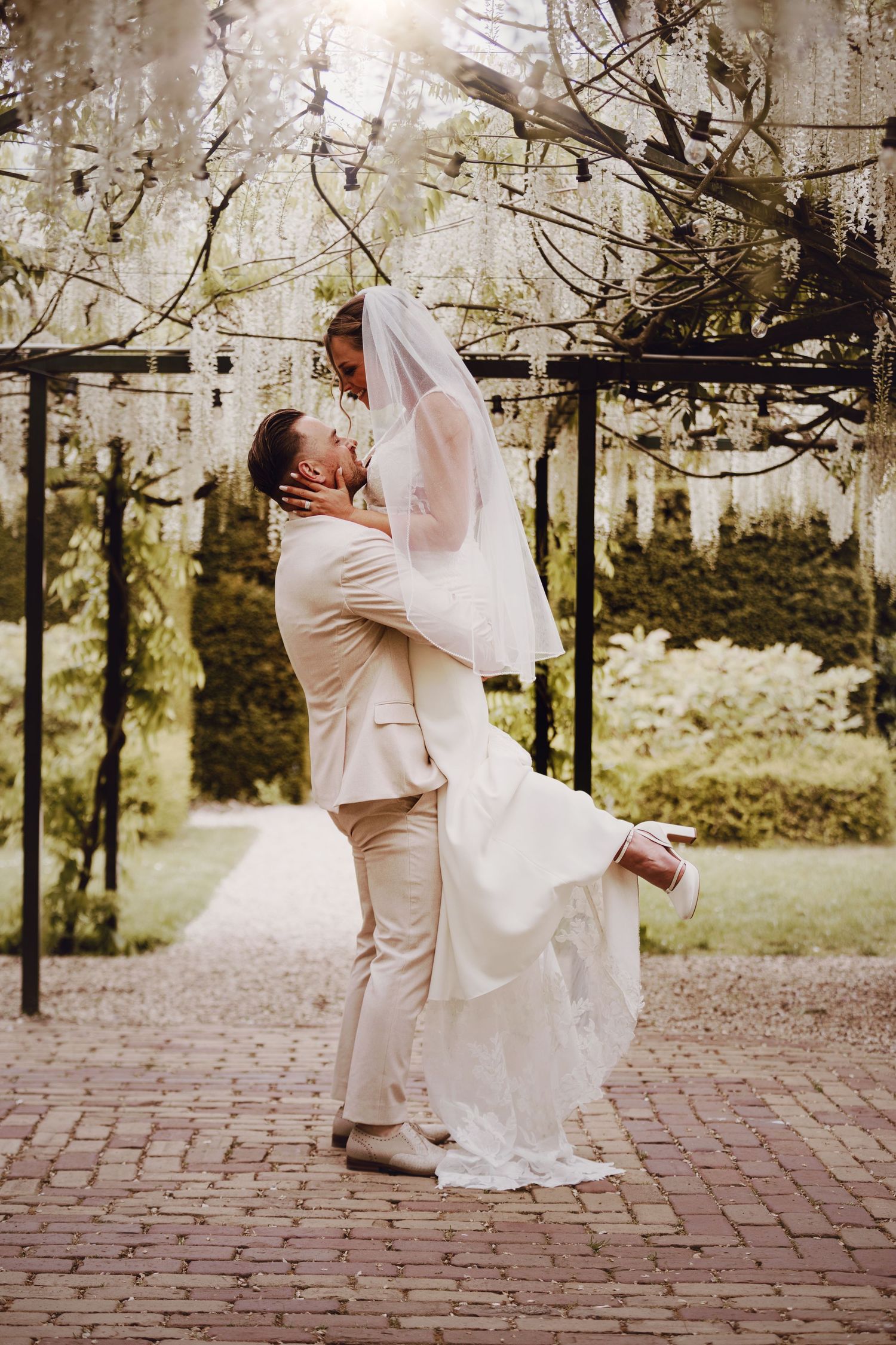 Groom lifts bride in white lace gown under blooming wisteria pergola in sunlit garden.