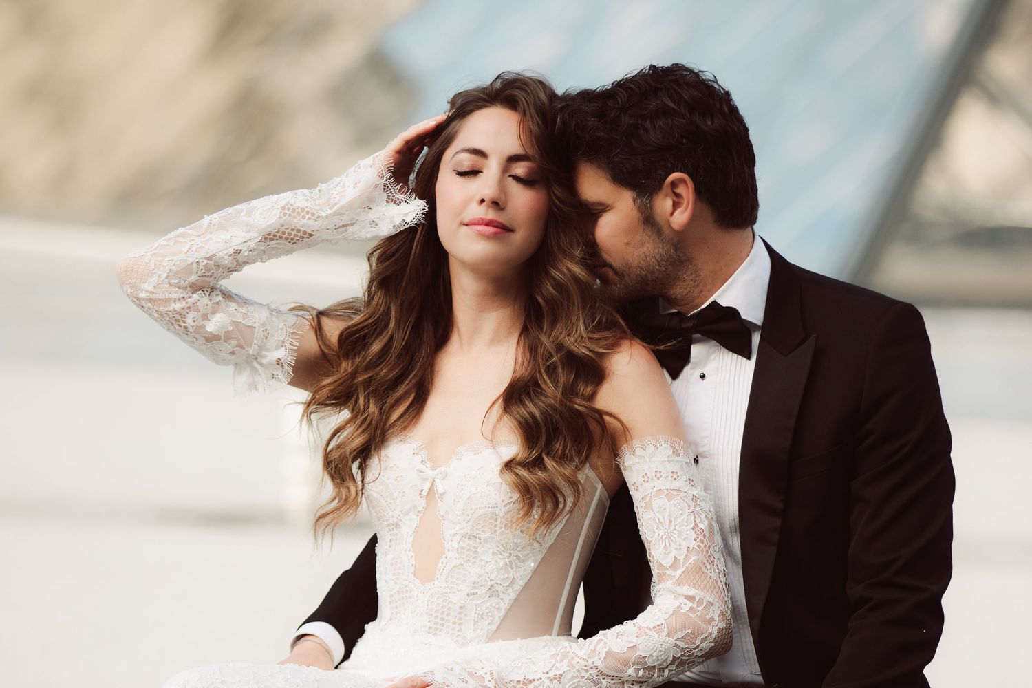 Elegant bride in lace gown with groom in tuxedo sharing an intimate moment near glass pyramid.