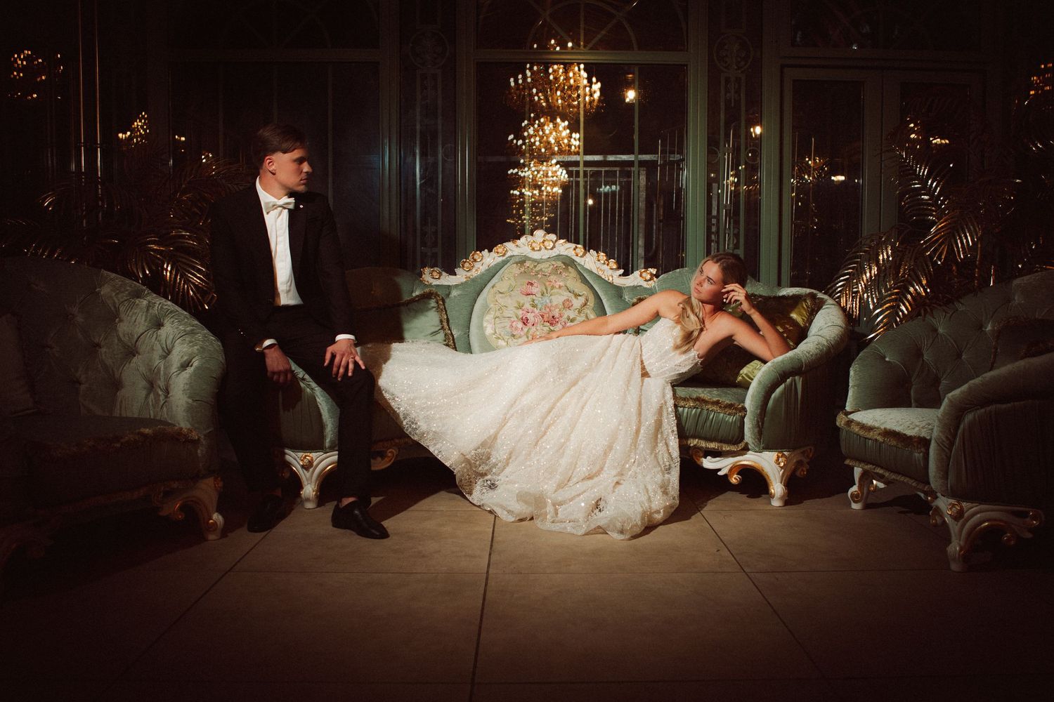 Elegant bride in white gown reclines on vintage green sofa beside groom in tuxedo in opulent chandelier-lit ballroom.
