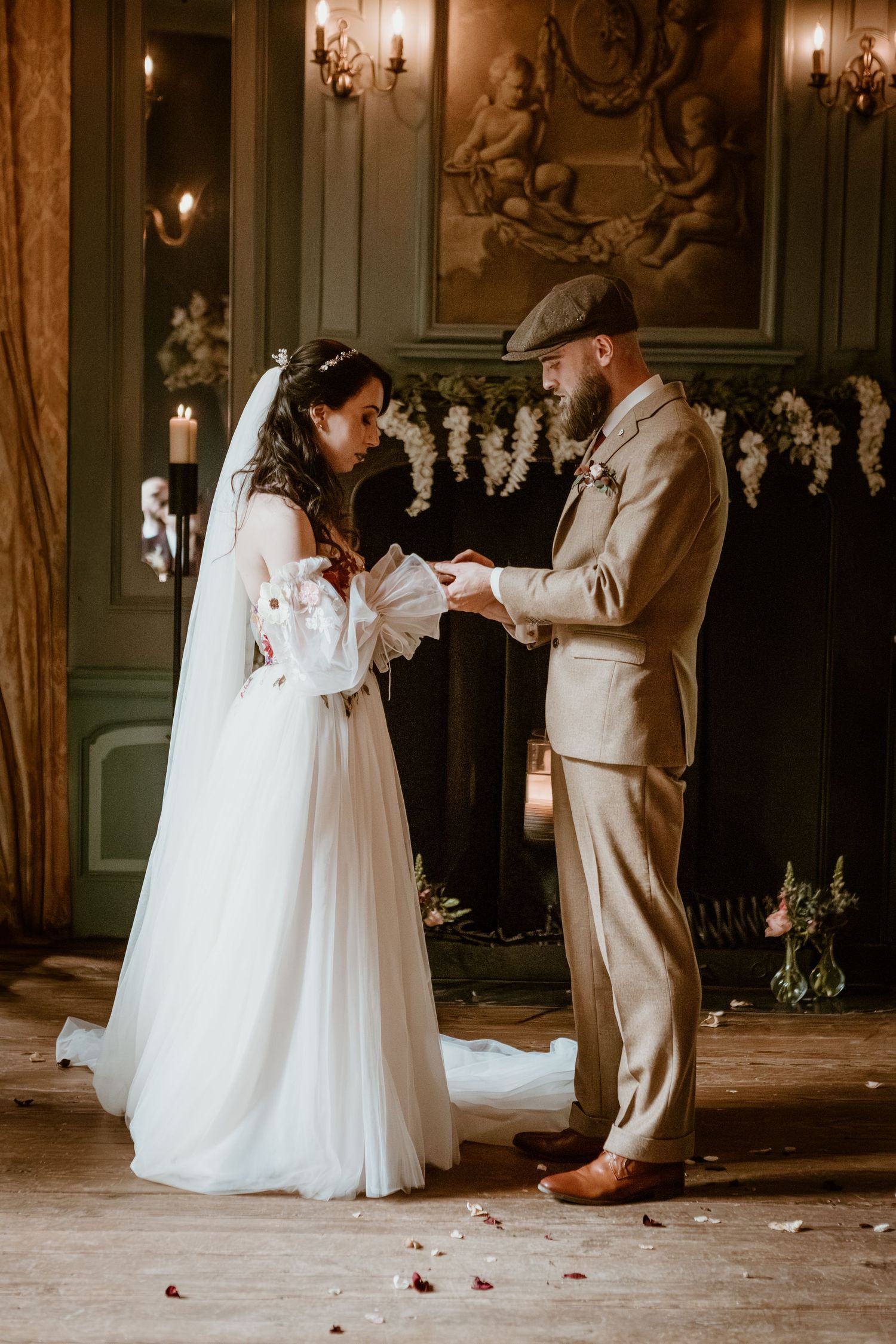 Bride in floral wedding gown and veiled groom exchange vows by candlelit fireplace.