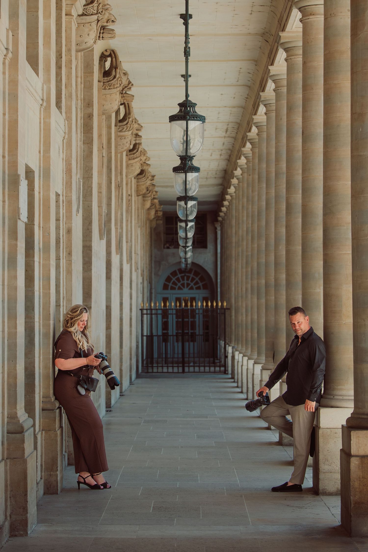 Two photographers review their cameras in a grand classical colonnade with ornate hanging lanterns.