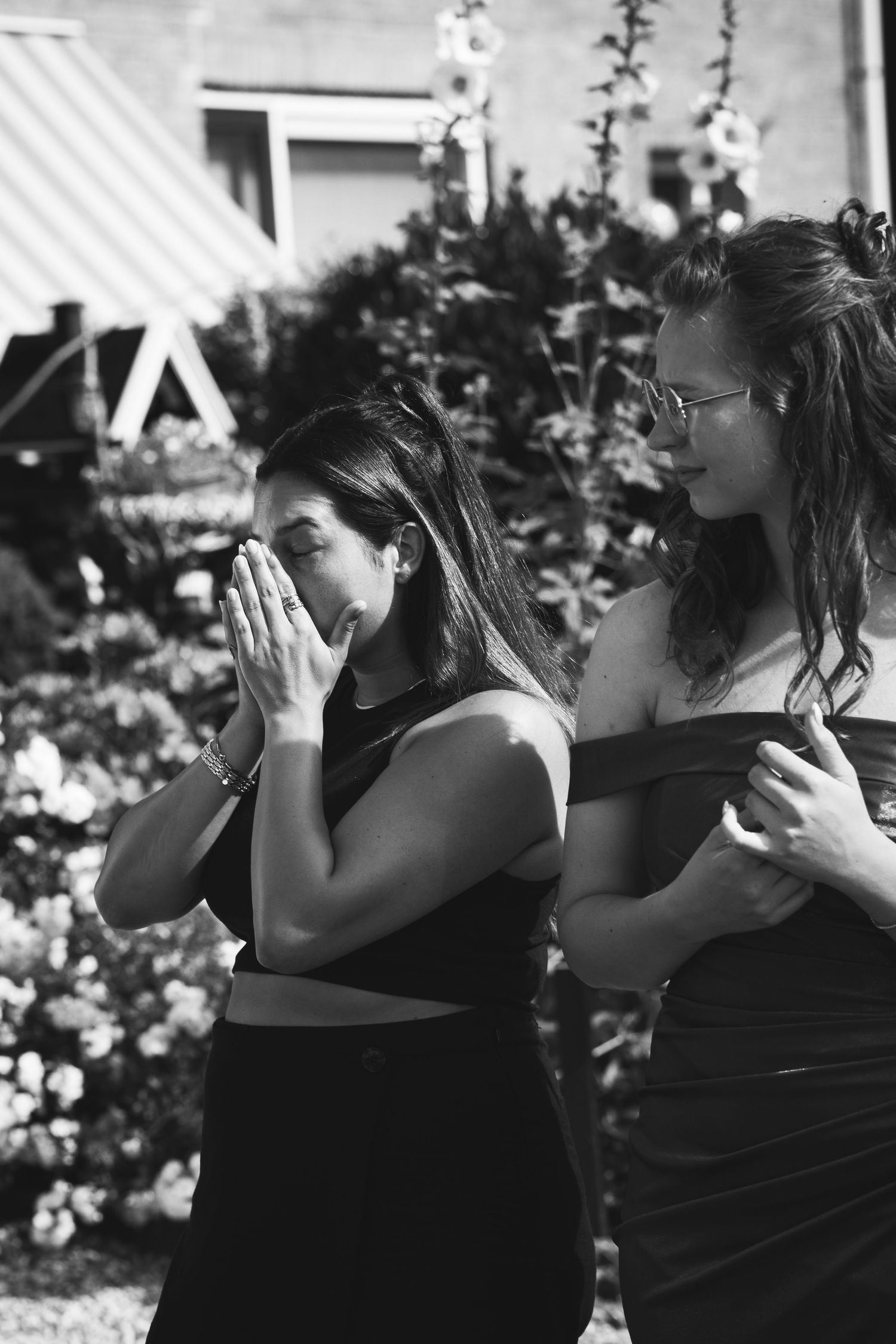 Two women in black dresses embrace outdoors in a garden setting.