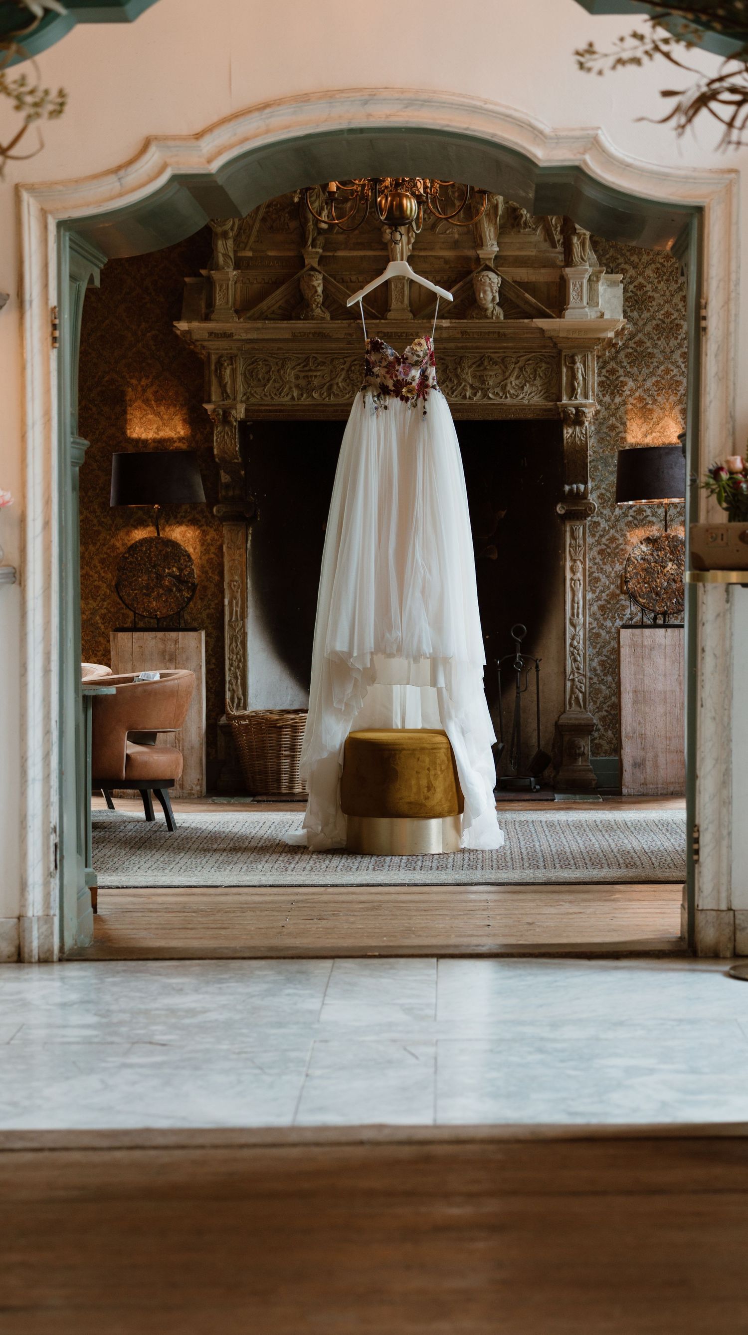 Elegant white wedding dress hanging in ornate vintage room with decorative fireplace.