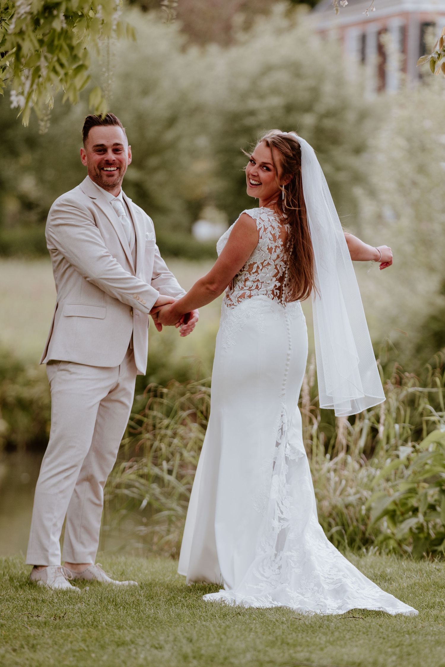 Smiling bride and groom in beige suit hold hands on green lawn near pond.